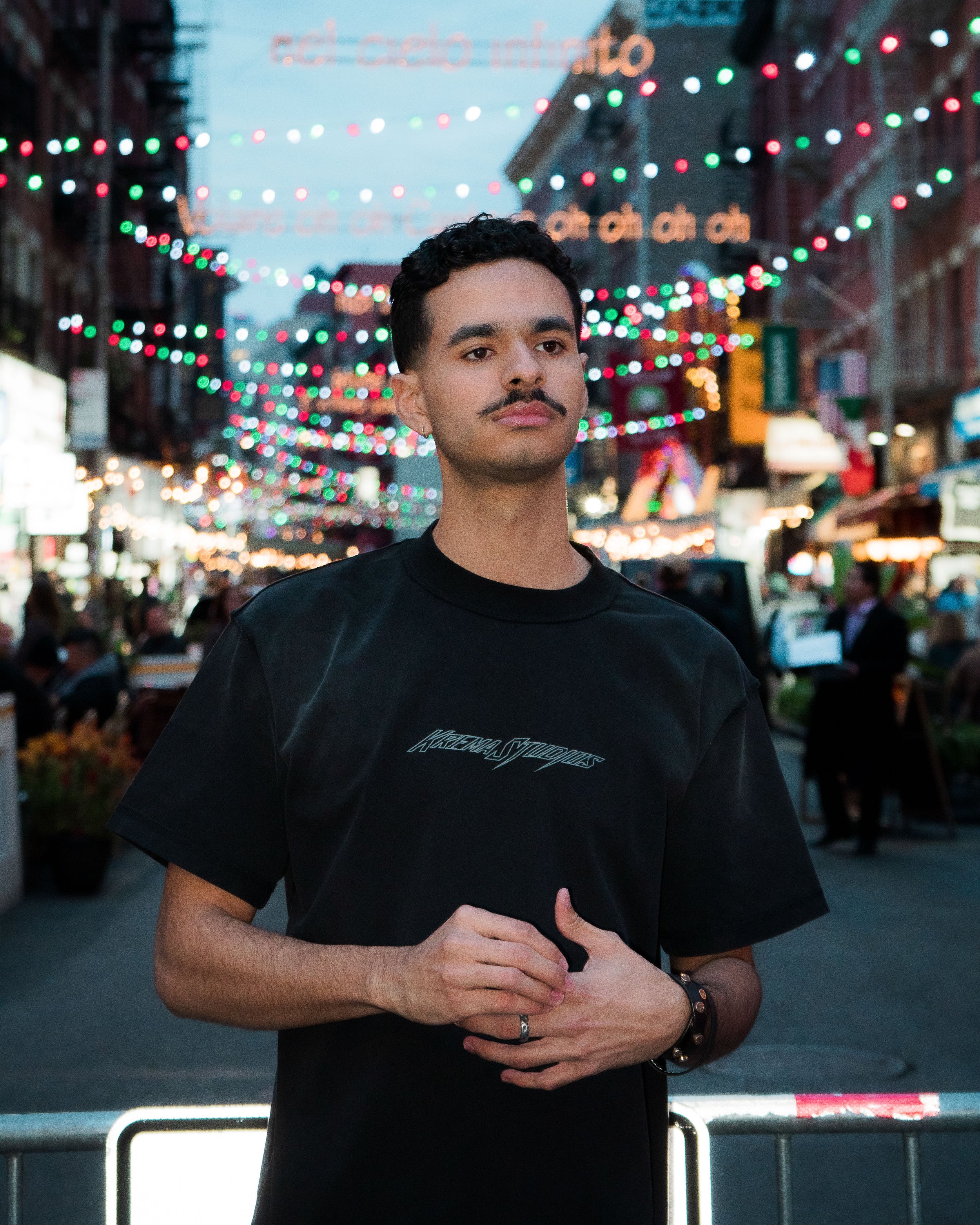 A young man with a mustache and short curly hair standing on a busy street at night, decorated with colorful string lights and blurred signage in the background.