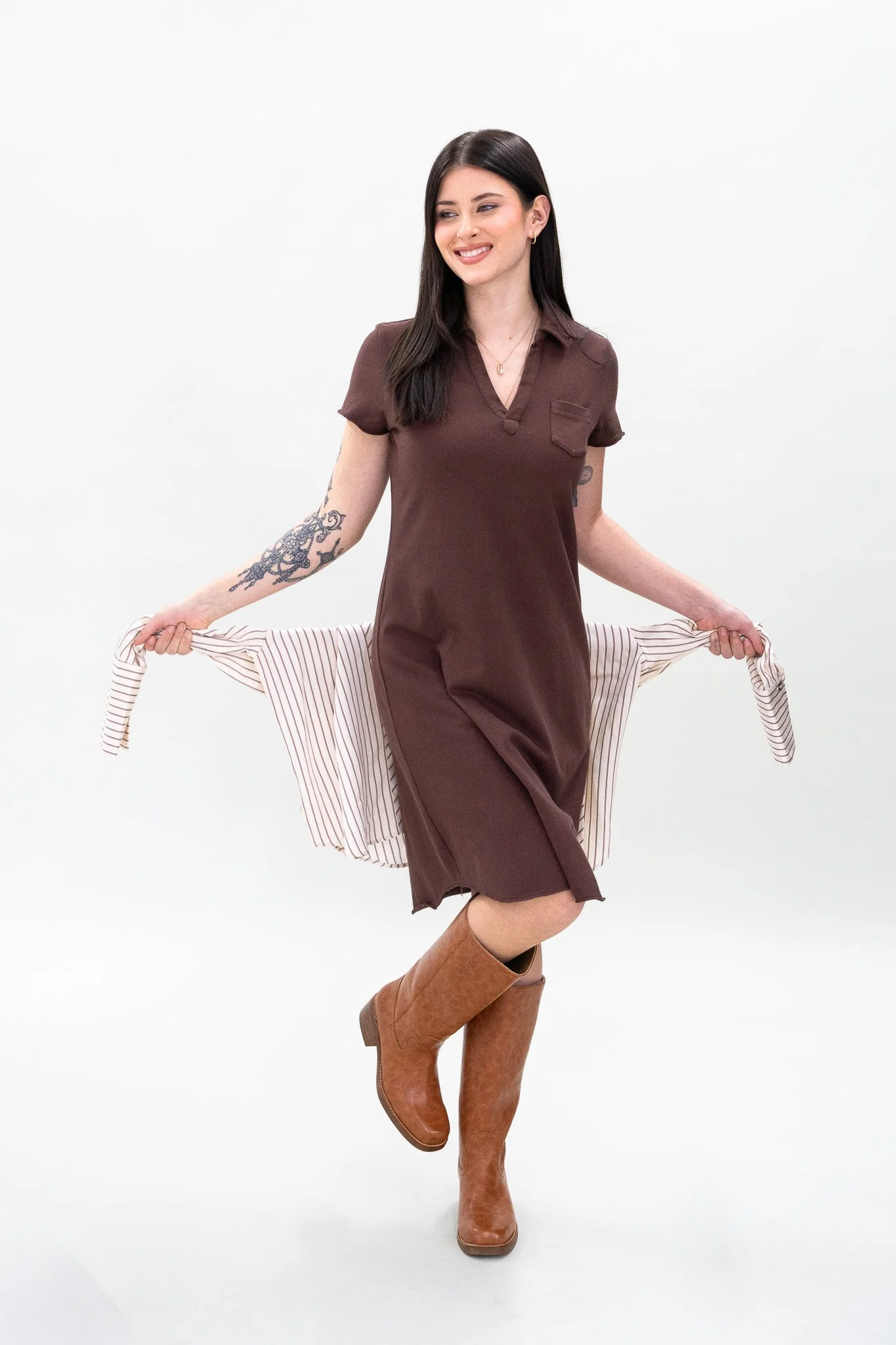 A woman with long dark hair and tattoos on her arms is smiling and holding a striped shirt behind her with both hands. She is wearing a brown dress and brown knee-high boots, standing against a plain white background.