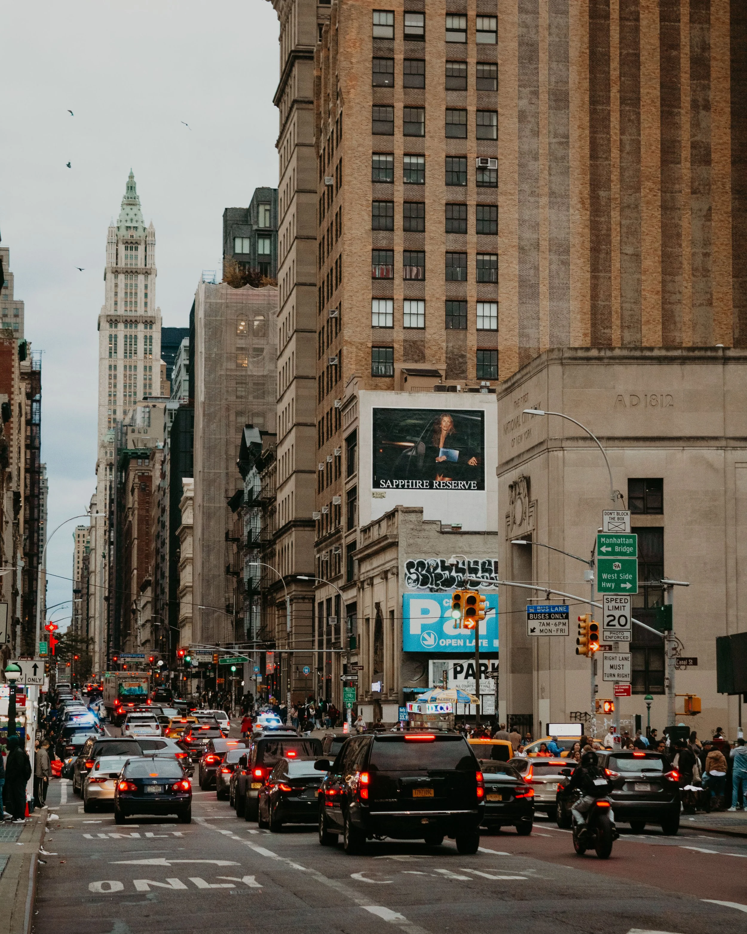 Busy city street with traffic, pedestrians, and tall buildings, including one with a digital billboard showing a woman with the text ''Sapphire Reserve.''