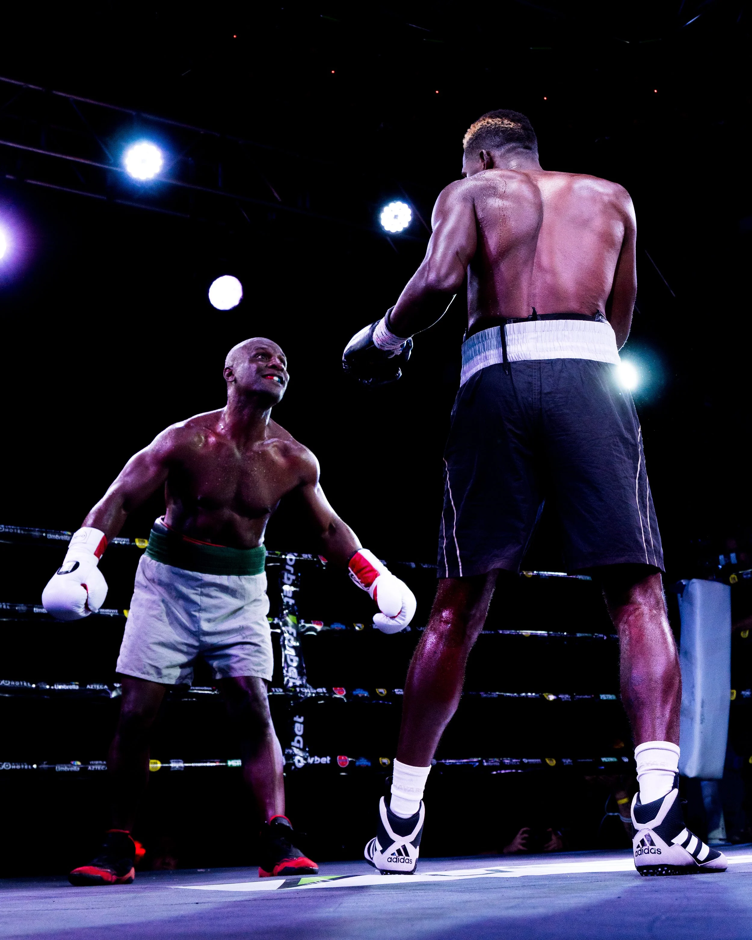 Two male boxers in a boxing ring, facing each other. One boxer has a muscular build and is seen from the back, wearing black shorts, white socks, and Adidas sneakers. The other boxer, who is bald, has a darker skin tone and a leaner physique, is wear