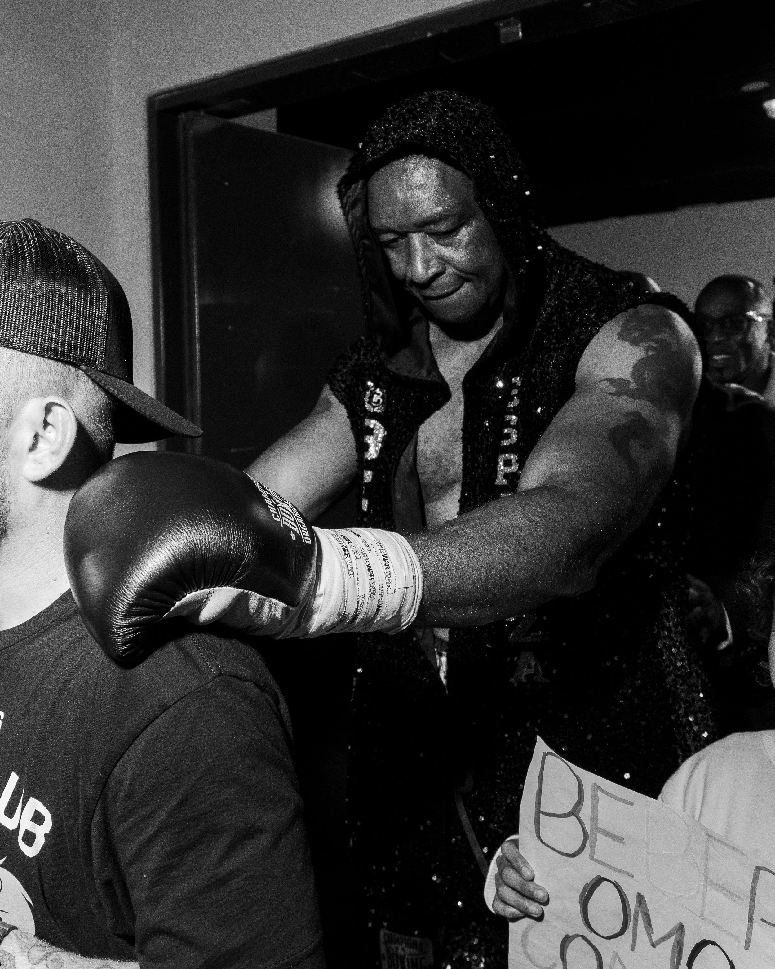Black and white photo of a man with boxers and boxing gloves in a gym, with a woman holding a sign that reads 'BE' and 'OM' in the foreground.