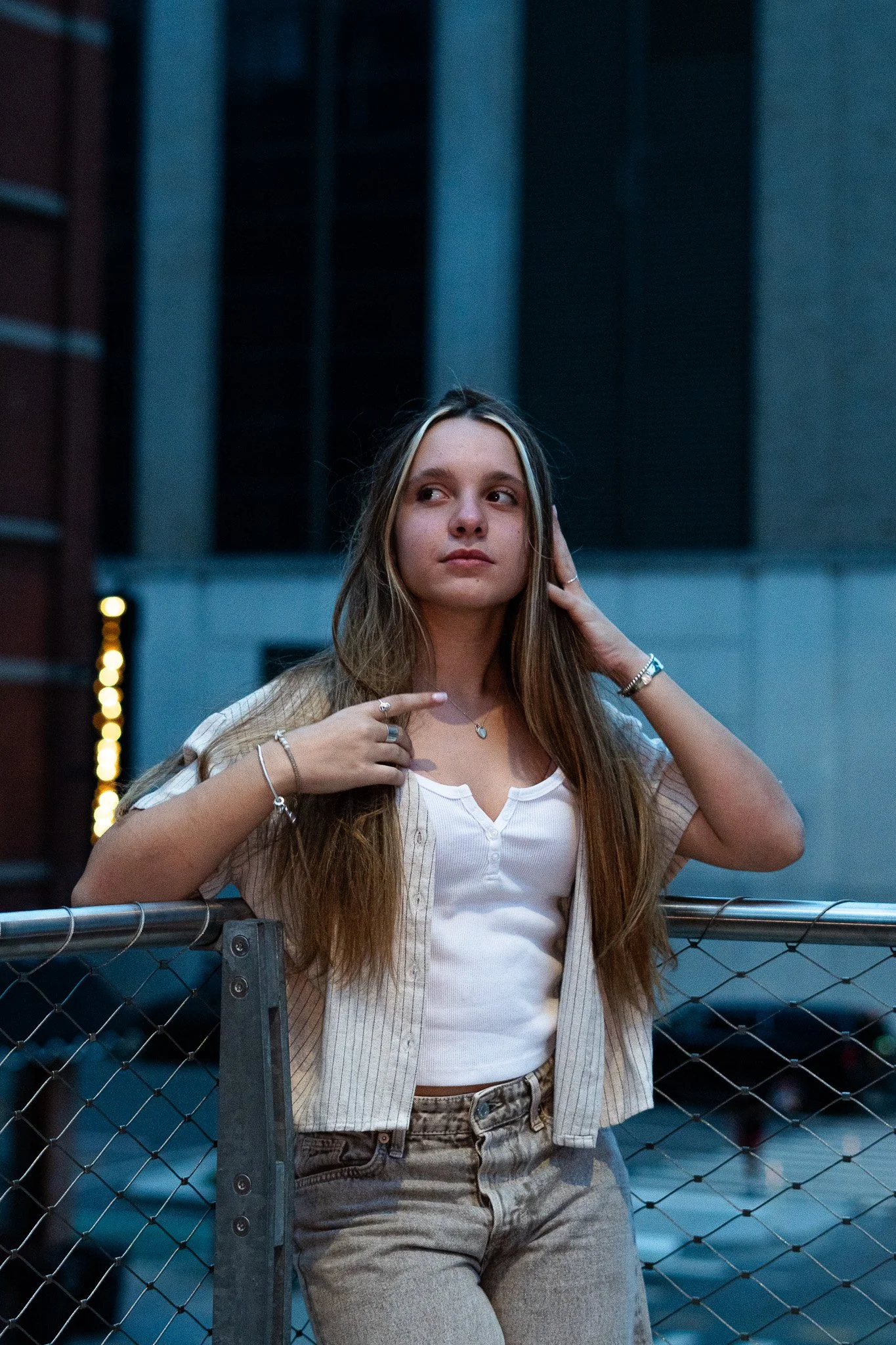 A young woman with long light brown hair standing outdoors near a chain-link fence, with a building and dark windows in the background, during dusk or evening. She is wearing a beige and white striped short-sleeve shirt, high-waisted jeans, and jewel