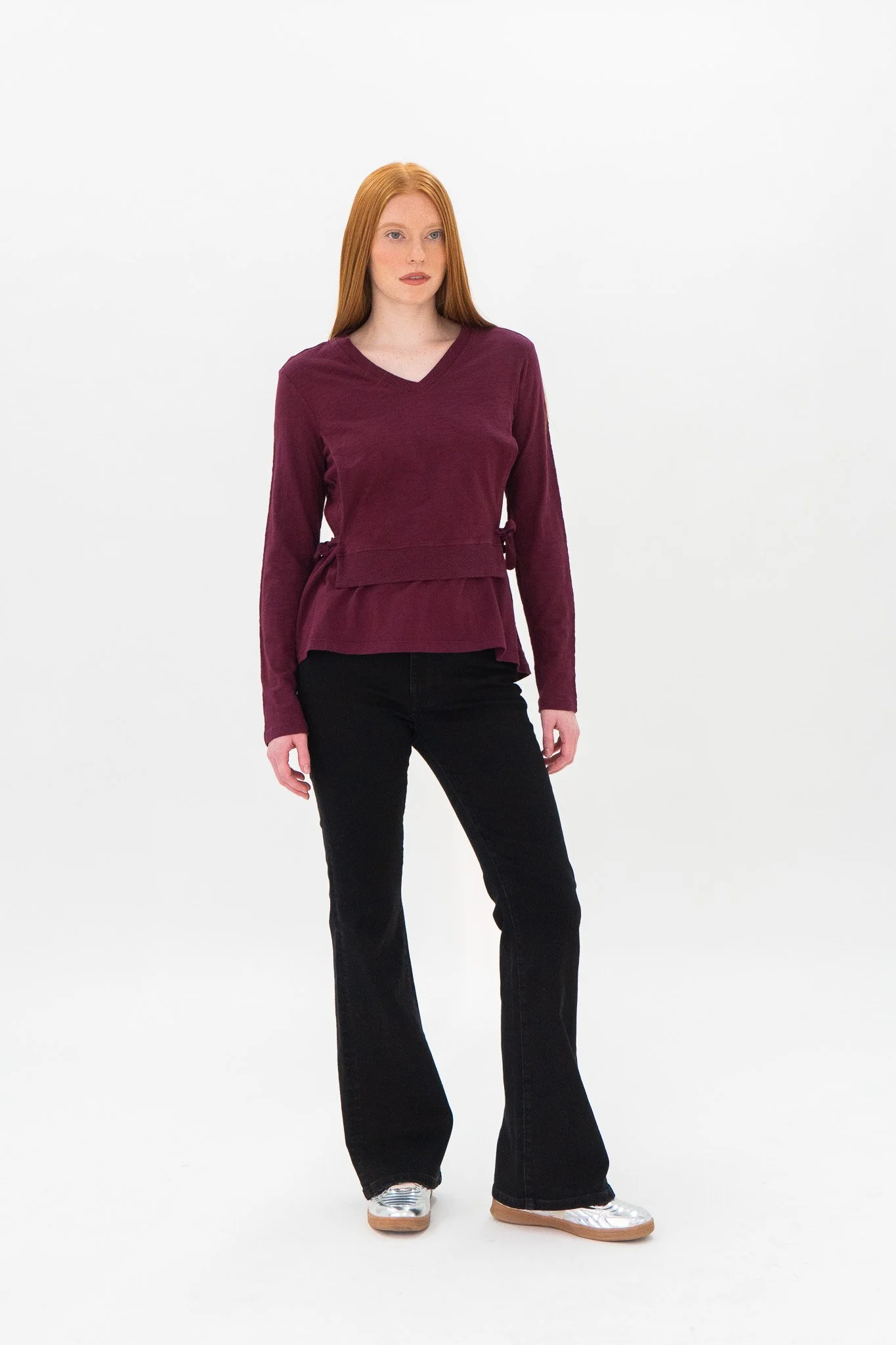 a woman with long red hair wearing a maroon long-sleeve top, black wide-leg pants, and silver sneakers, standing against a plain white background