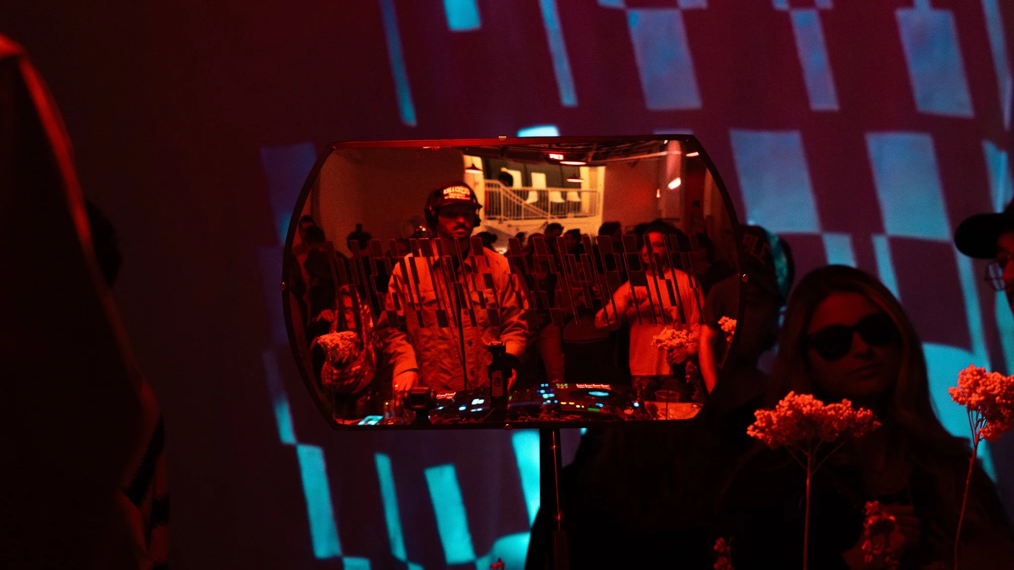 Reflection of a DJ in a mirror at a party, with people in the background and red and blue lighting.