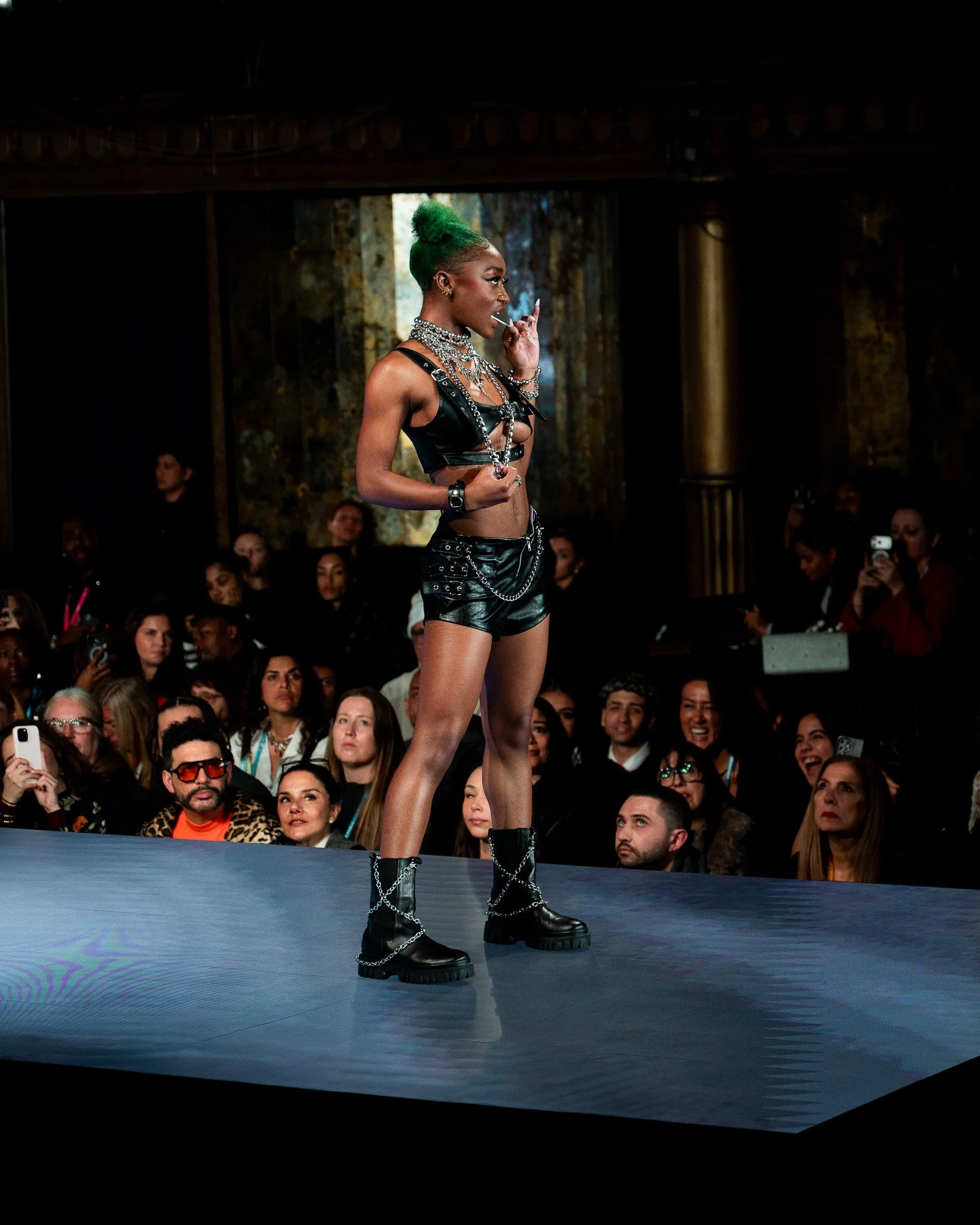 A woman with green hair in a high bun, wearing black leather clothing and chunky black boots with chains, stands on a runway at a fashion show. She is surrounded by an audience, some taking photos.