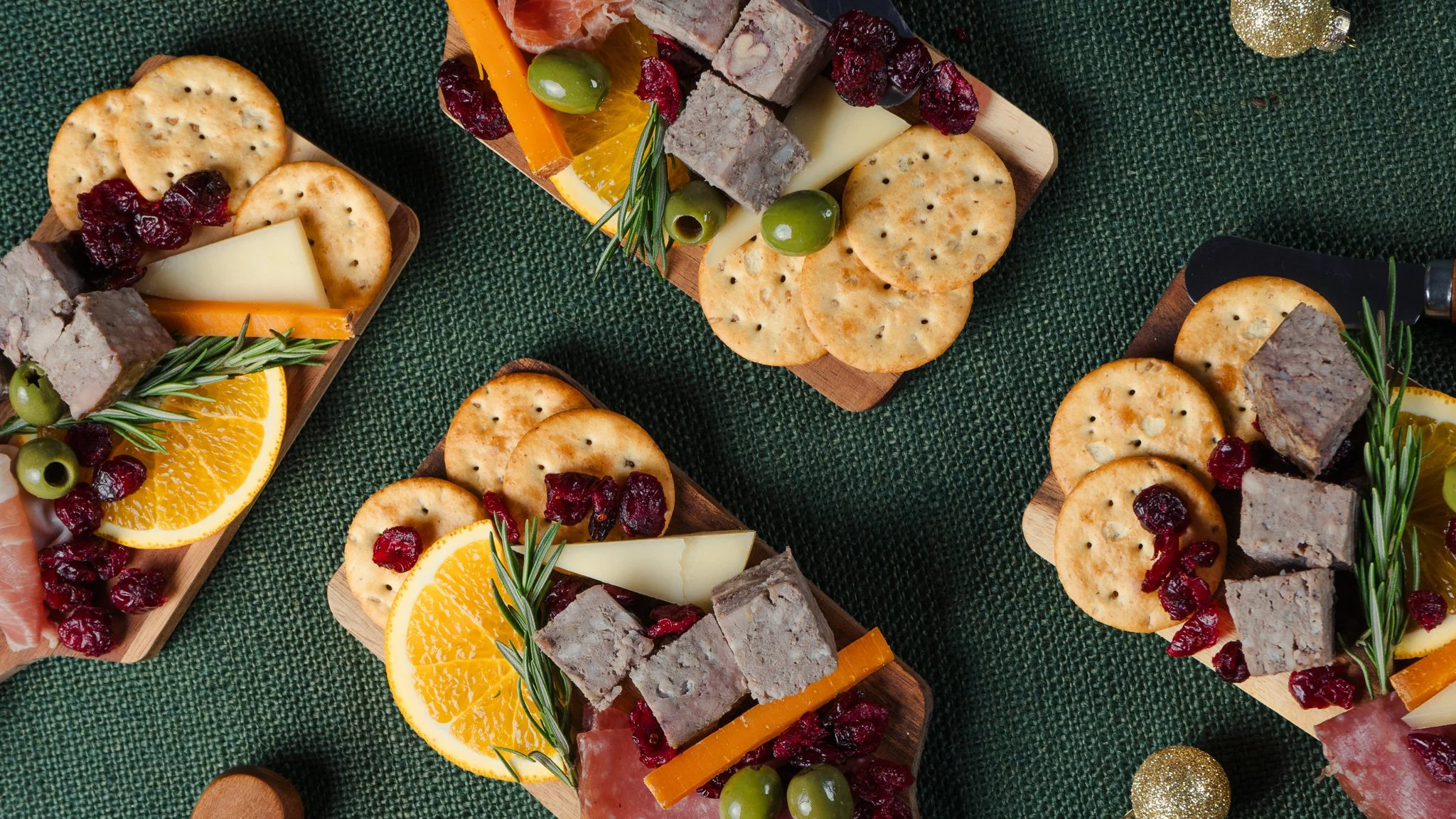 Assorted cheese and cracker platter with sliced oranges, green olives, sprigs of rosemary, and decorative gold ornaments, arranged on wooden trays on a green textured surface.