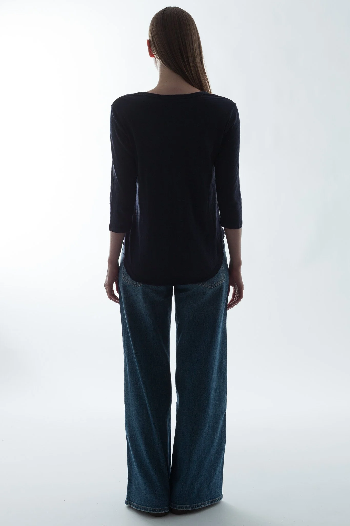 Back view of a woman with long brown hair wearing a black long-sleeve top and wide-leg blue jeans, standing against a plain, light background.