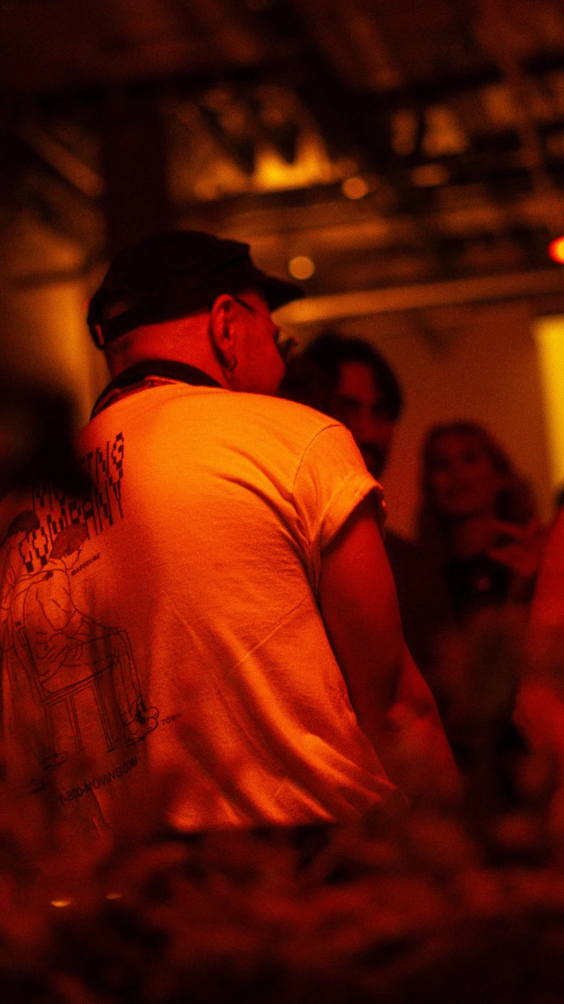 Person wearing glasses and a cap, seen from the side, in a dimly lit room with blurred background.