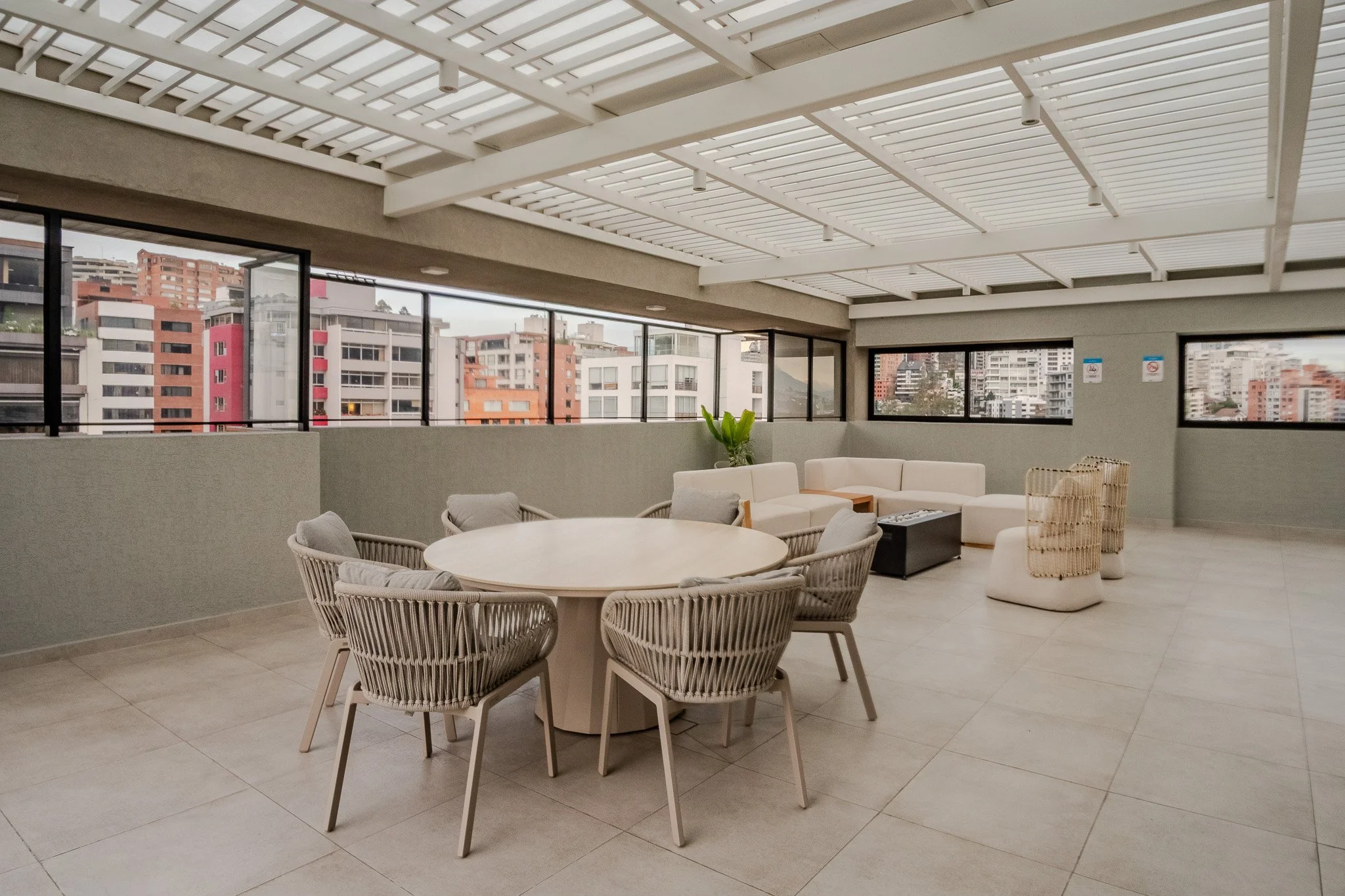 Indoor lounge area with round table, wicker chairs, white sofa, potted plant, large windows showing city buildings, white pergola ceiling.