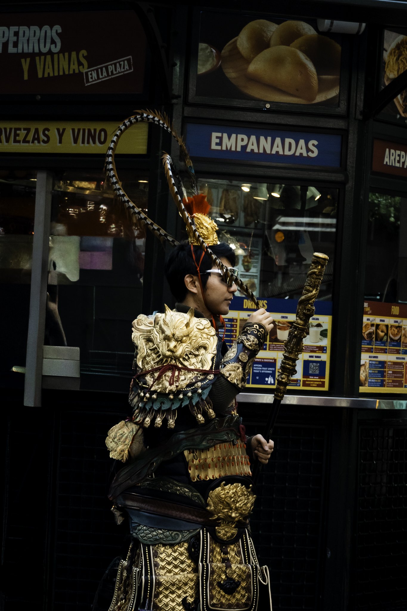 A person dressed in traditional Asian warrior costume with intricate gold and black armor, holding a sword, standing in front of a food stall with signage for empanadas and other foods.