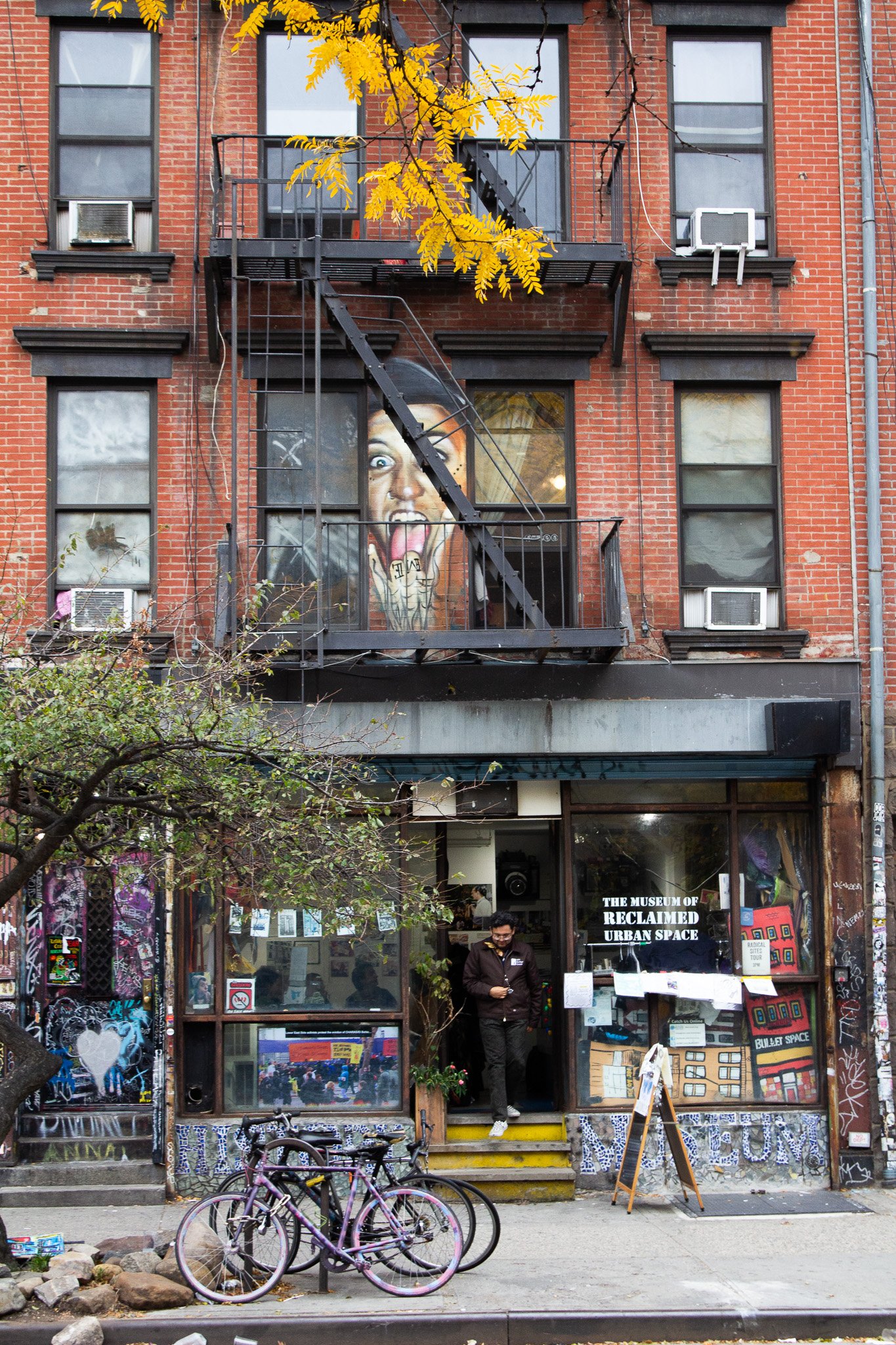 The exterior of a building with a large mural of a woman screaming and sticking out her tongue on the second floor, a storefront labeled "The Museum of Reclaimed Urban Space" on the ground level, several bicycles parked outside, and a person exiting 