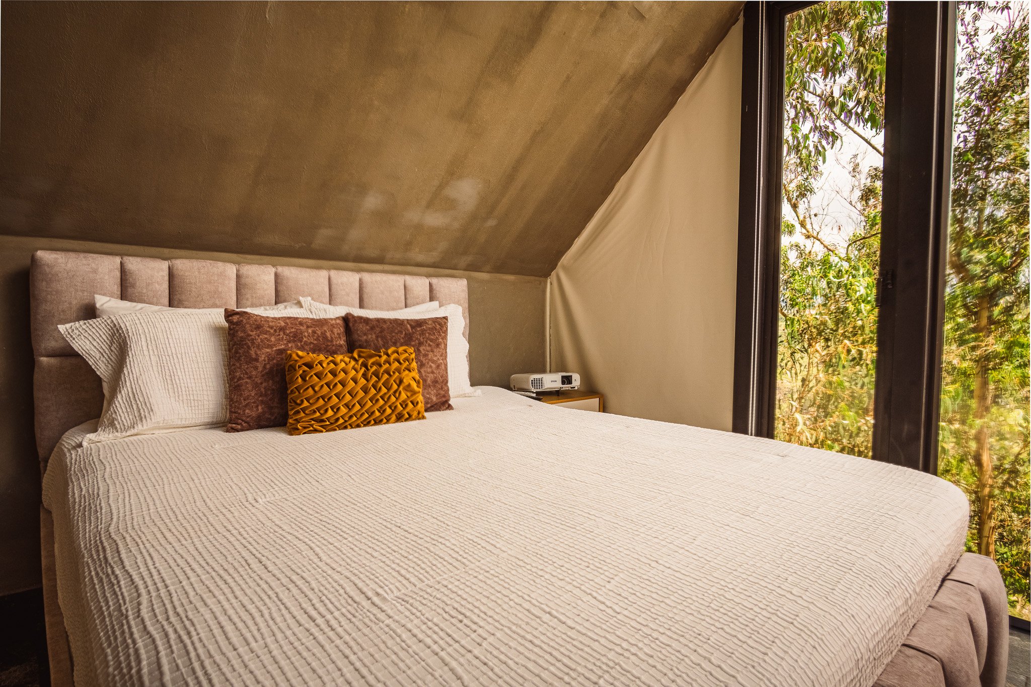 A cozy bedroom with a beige upholstered bed featuring white and brown pillows, situated under a slanted ceiling. The window shows green trees outside, and a small nightstand with a projector on it is beside the bed.