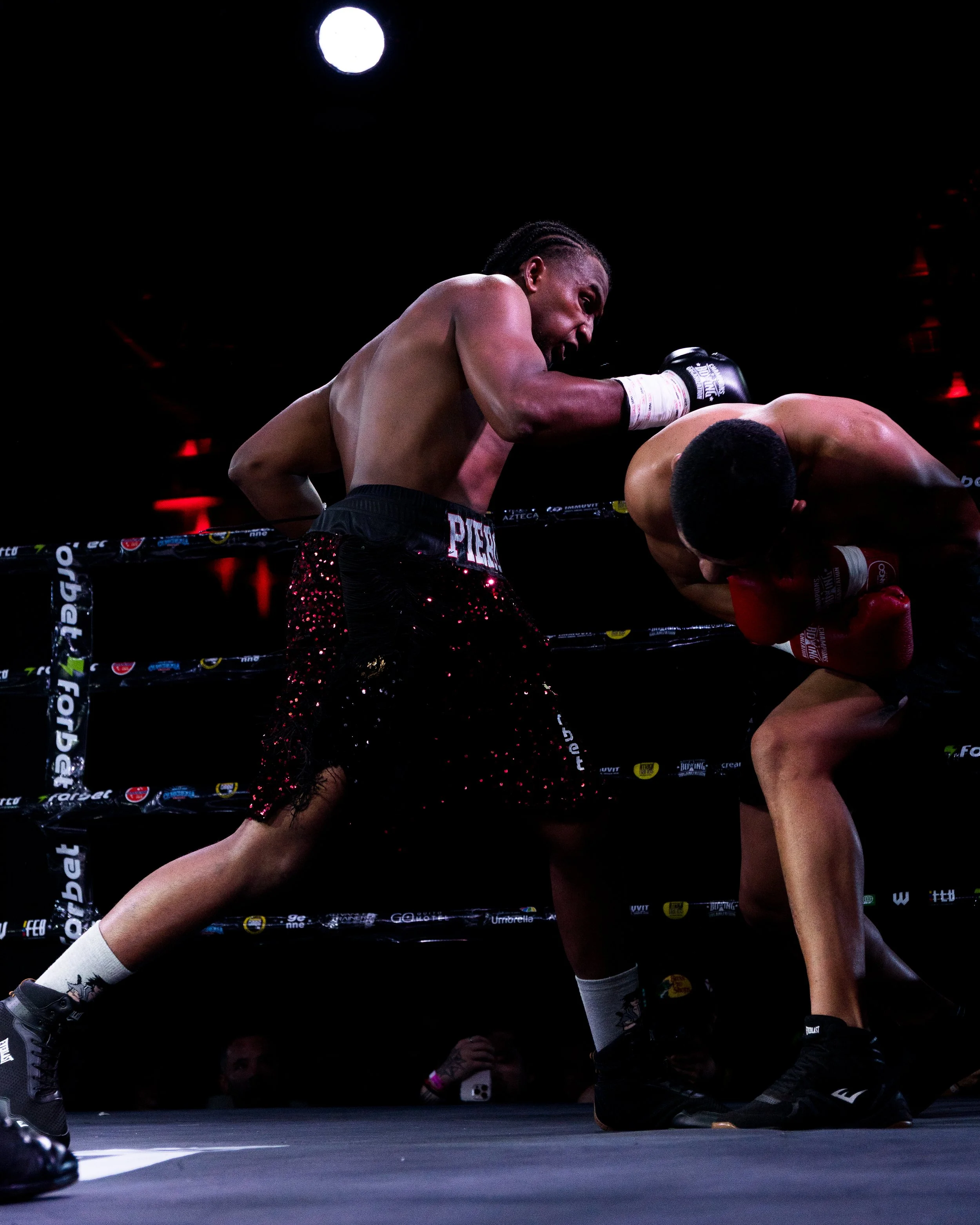 Two boxers fighting inside a ring at night, with one boxer leaning over the other as they engage in a match.