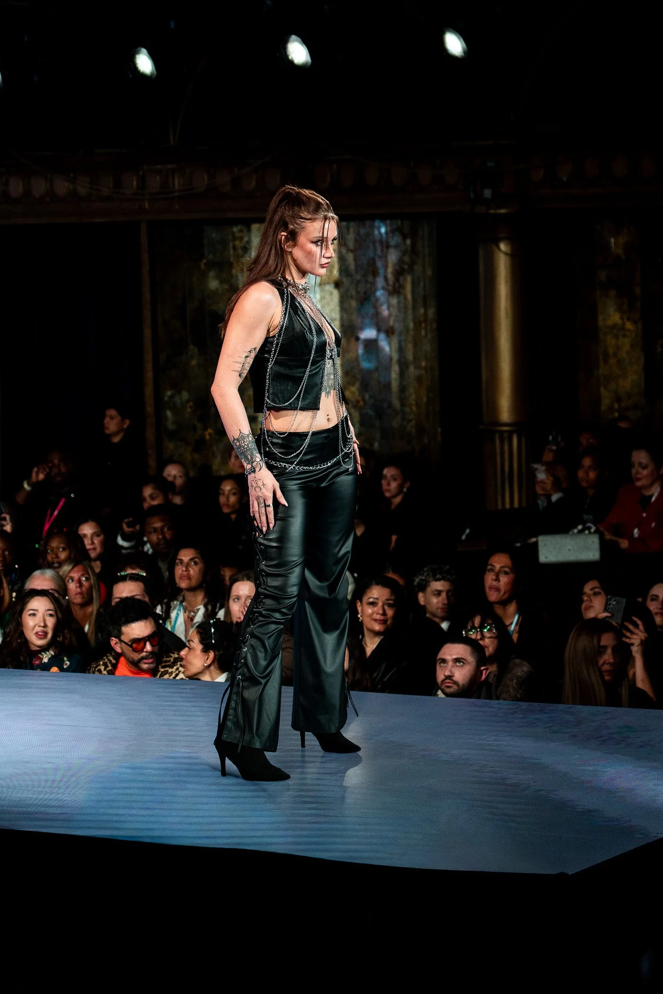 A woman walking on a runway during a fashion show, dressed in black leather pants and vest with chains, with an audience in the background.