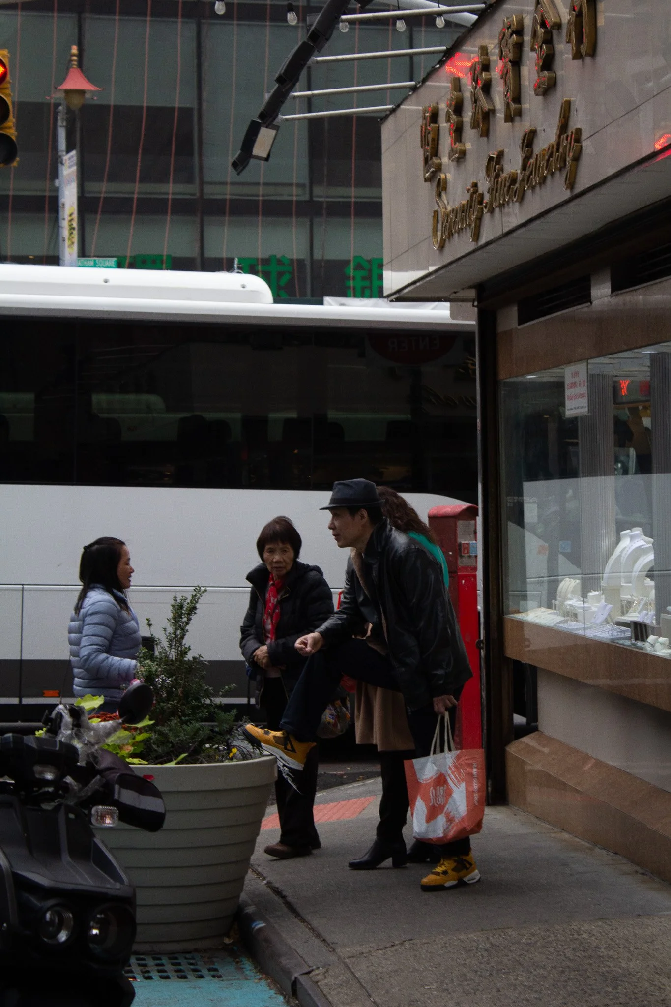 People walking on a city sidewalk near a jewelry store, with a bus and tall buildings in the background.