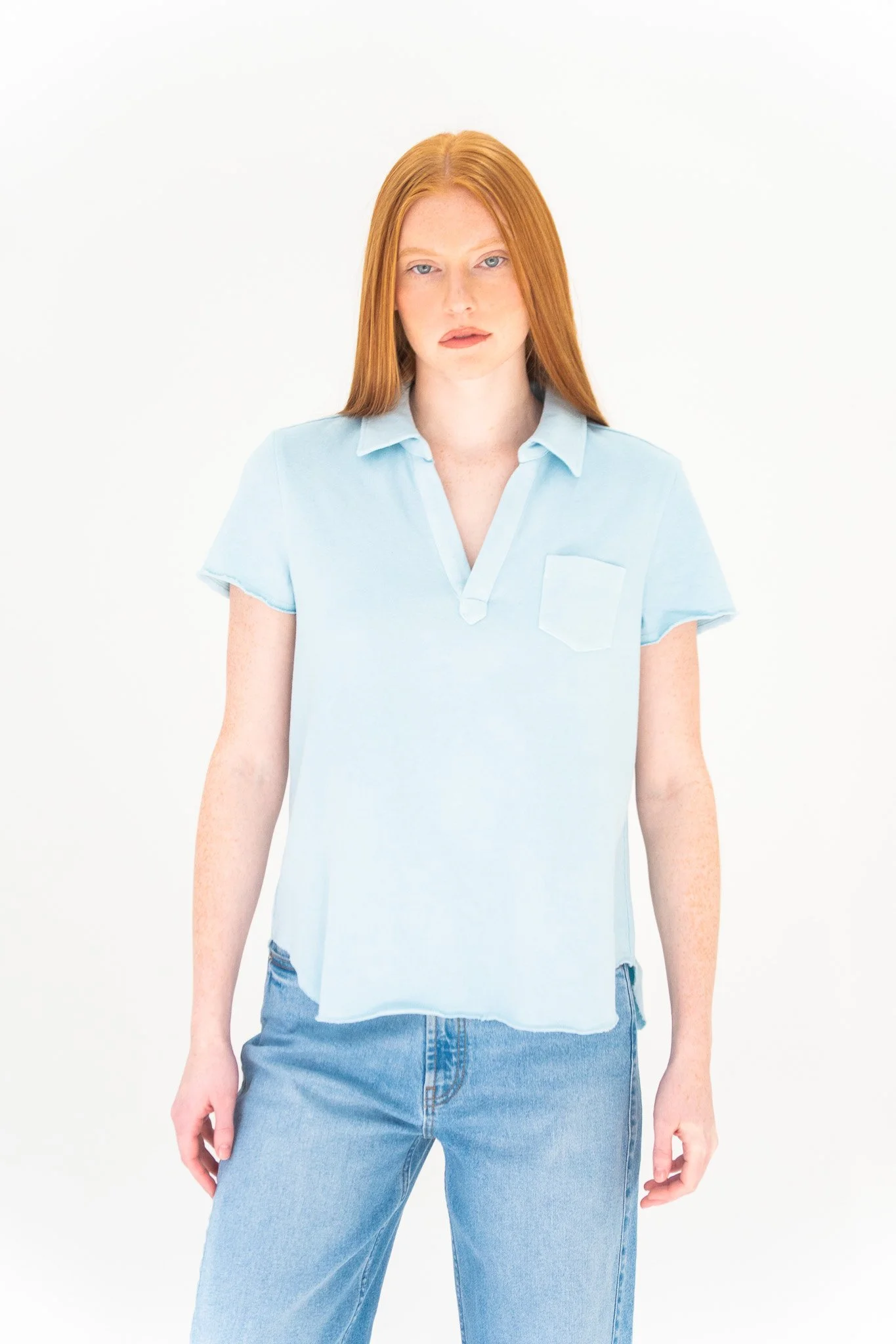 A woman with long red hair wearing a light blue short-sleeved button-up shirt and light blue jeans standing against a white background.