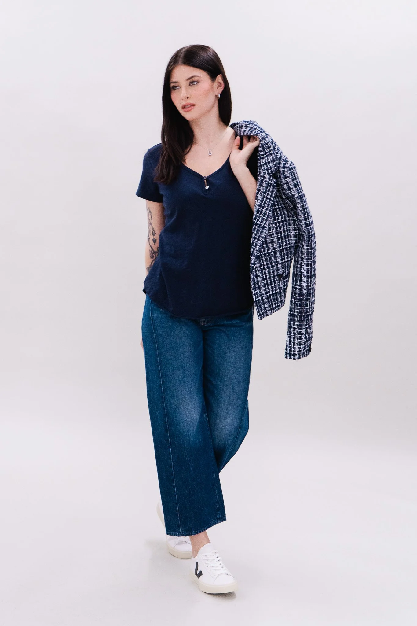 A woman with long dark hair, wearing a blue short-sleeve top, blue jeans, and white sneakers, stands against a plain white background. She carries a blue and white checkered blazer over her shoulder and looks to the side.