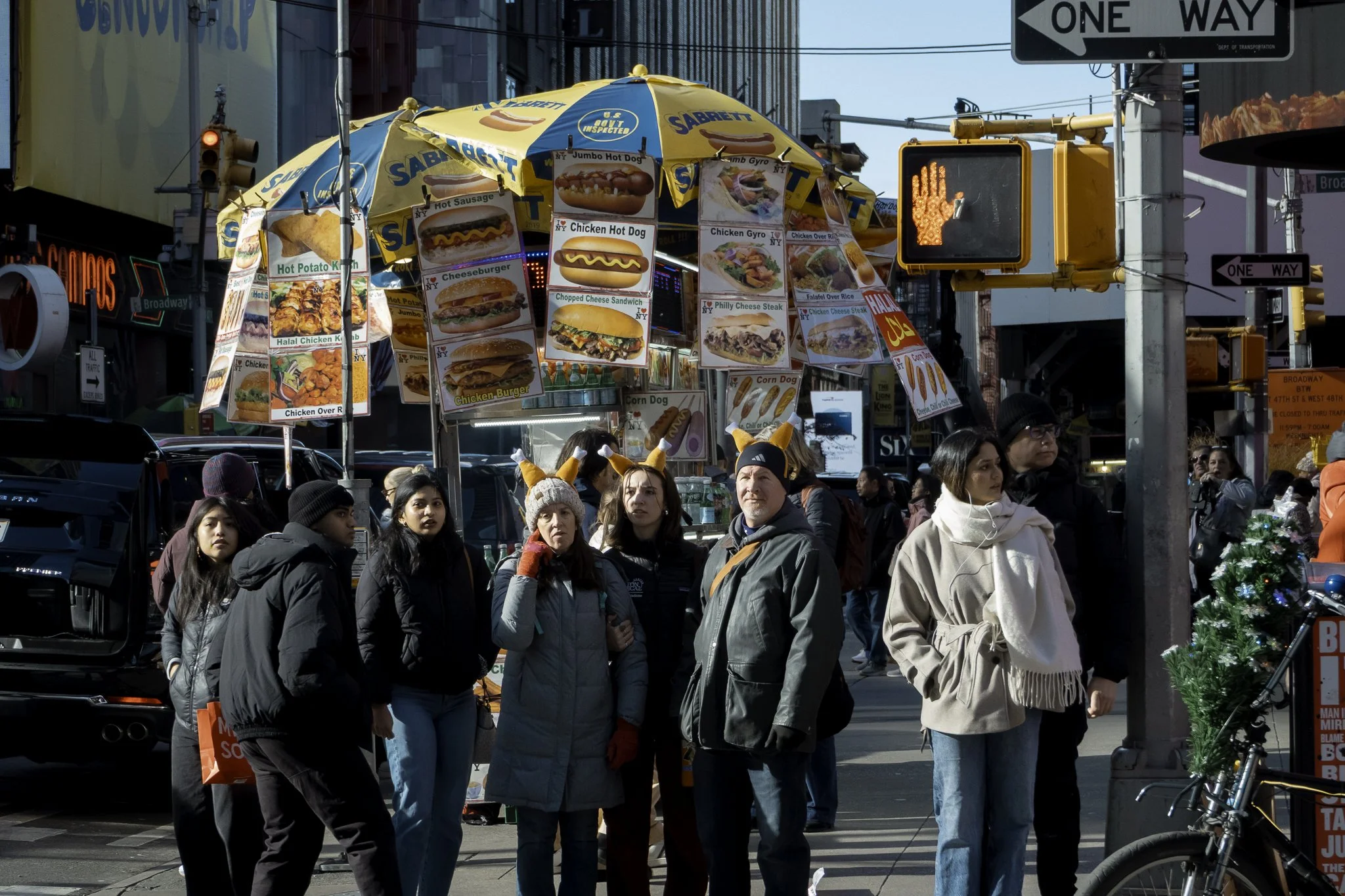 Crowd of people waiting at a street food stand with signs displaying various fast food options like hot dogs, cheeseburgers, chicken sandwiches, and fries, with traffic lights and street signs in the background.