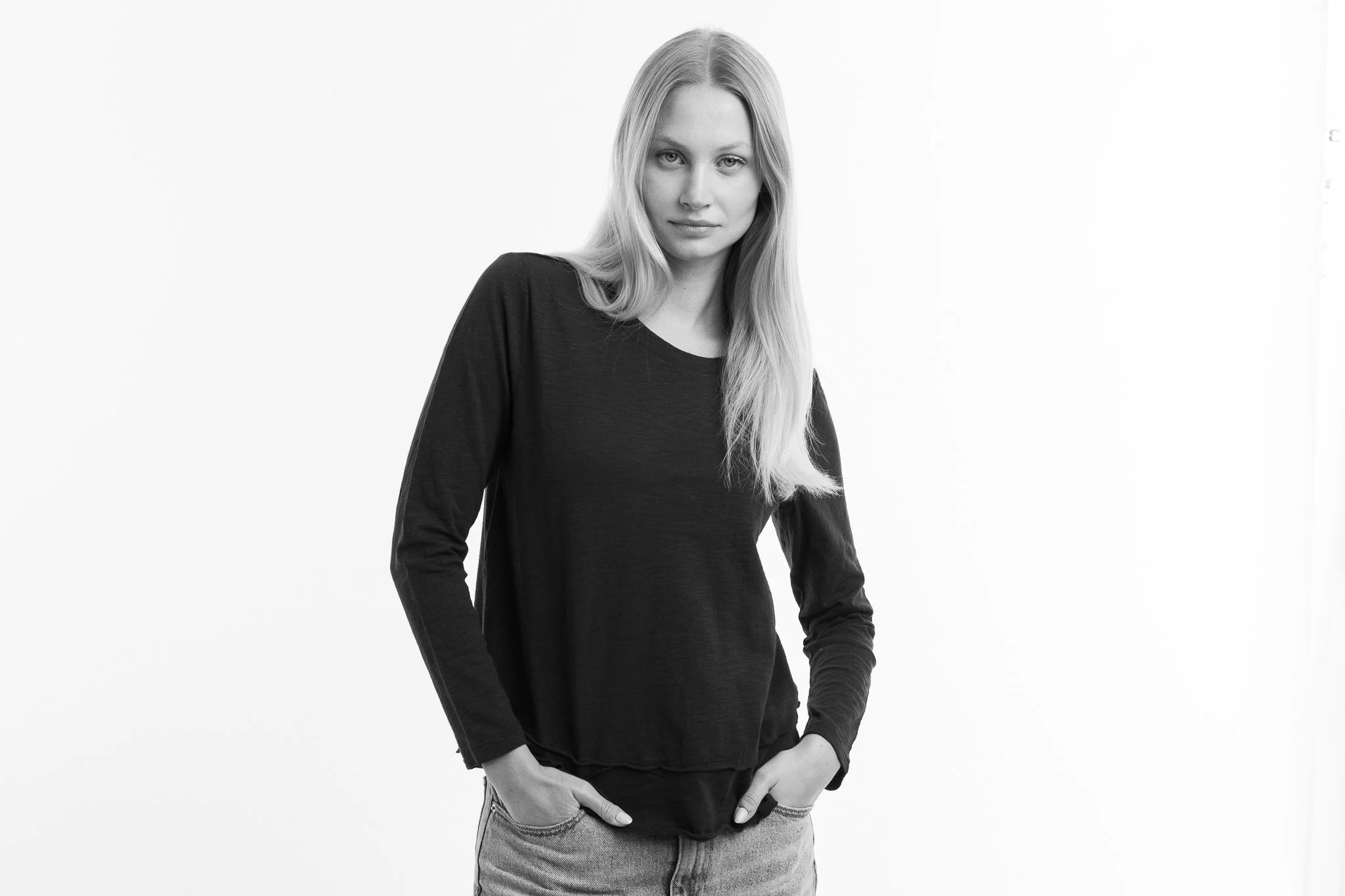 Black-and-white photo of a young woman with long hair, wearing a long-sleeved black top and light-colored jeans, standing against a plain background.