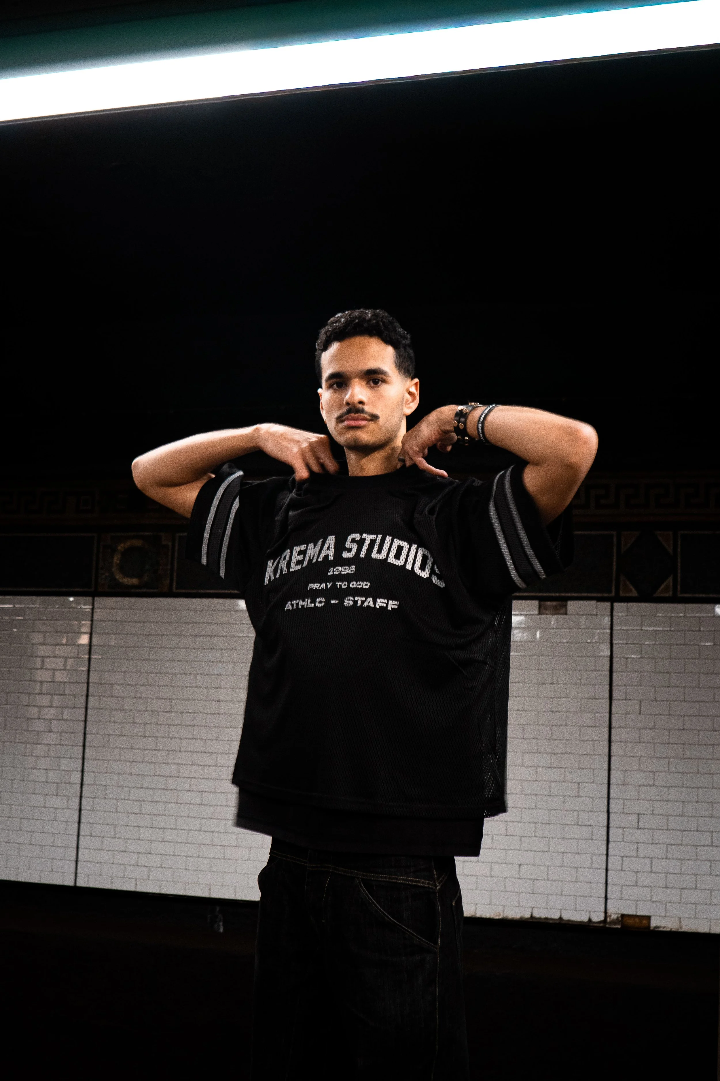 A young man in a black sports jersey standing in a subway station, adjusting his hair.
