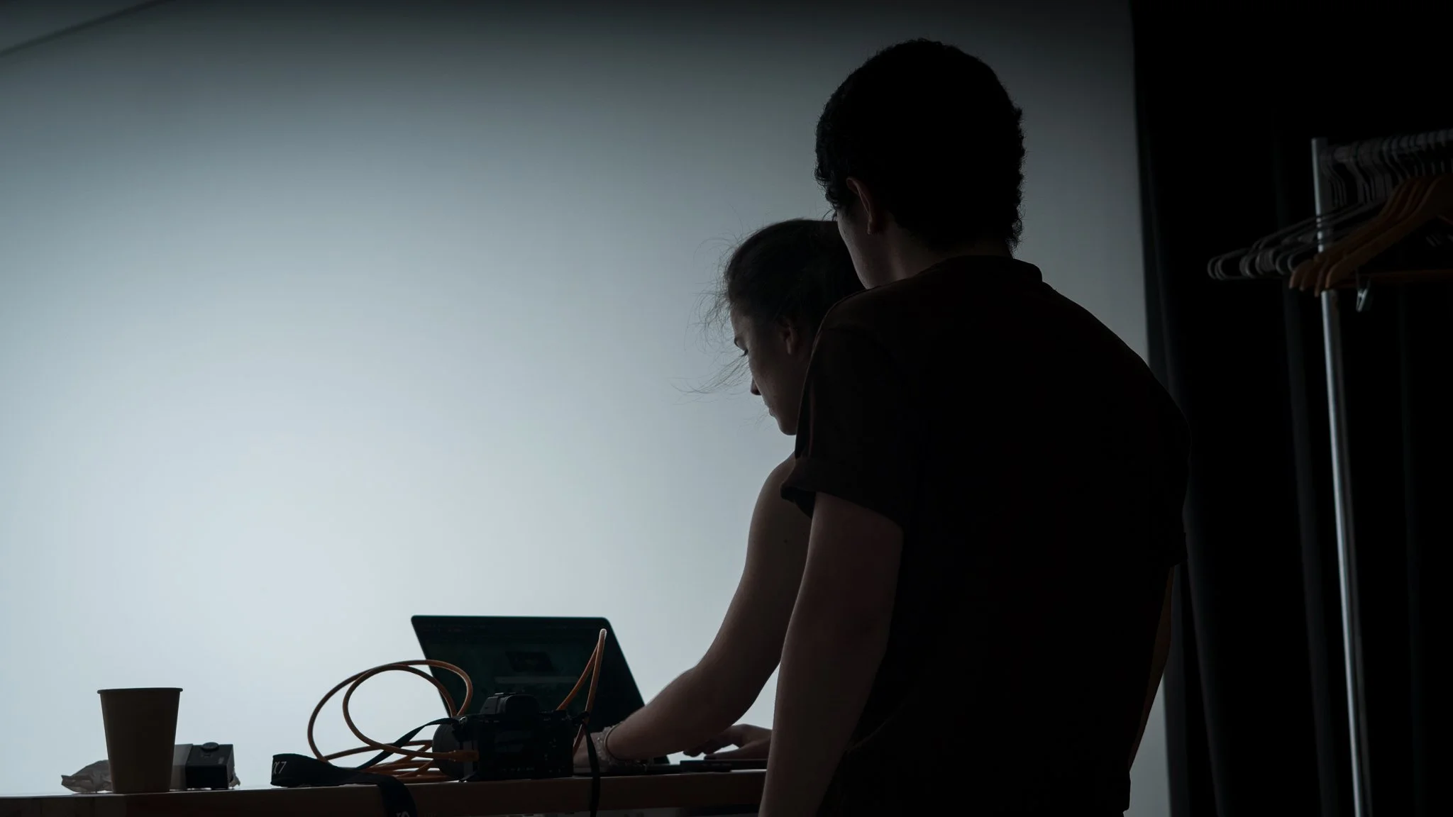 Silhouette of two people, one standing behind the other, in a room with a closet and a table with electronic equipment and a paper cup.