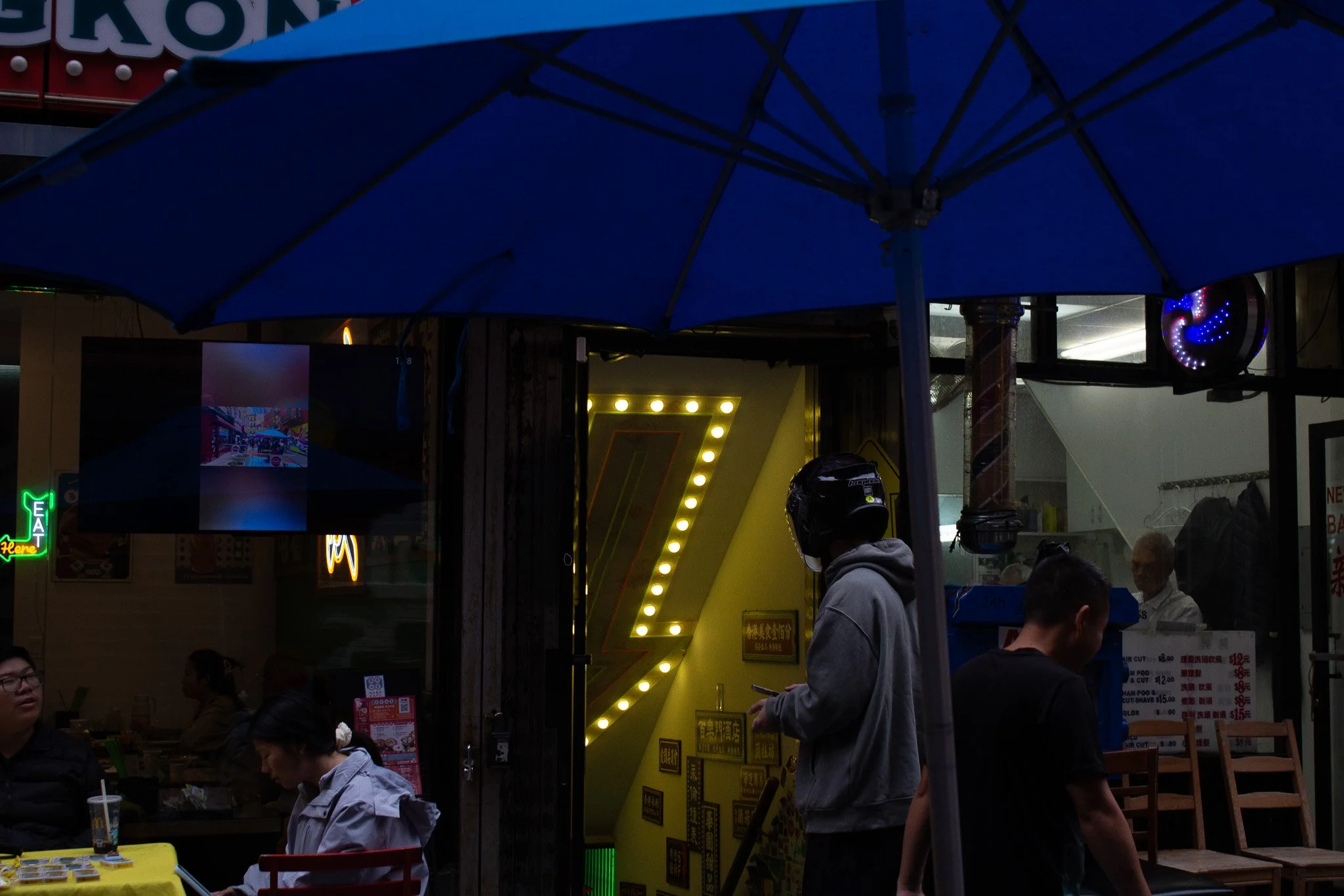 People dining outside a restaurant with neon signage, illuminated 'Z' sign, and a blue umbrella overhead.