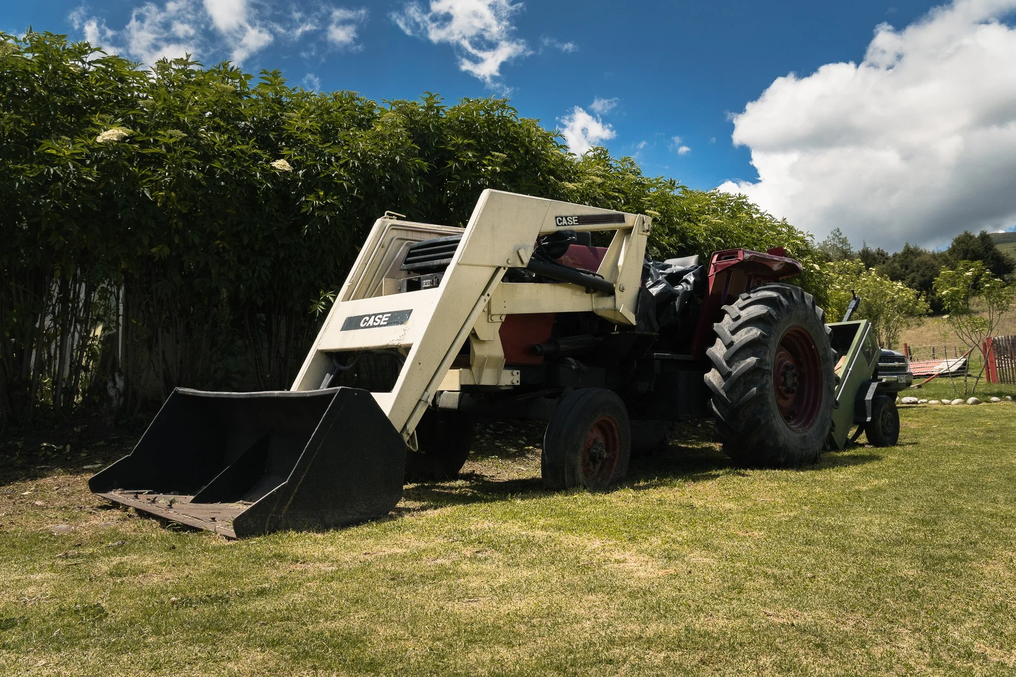 A tractor parked on a grassy area near a dense green hedge with a blue sky and scattered clouds overhead.