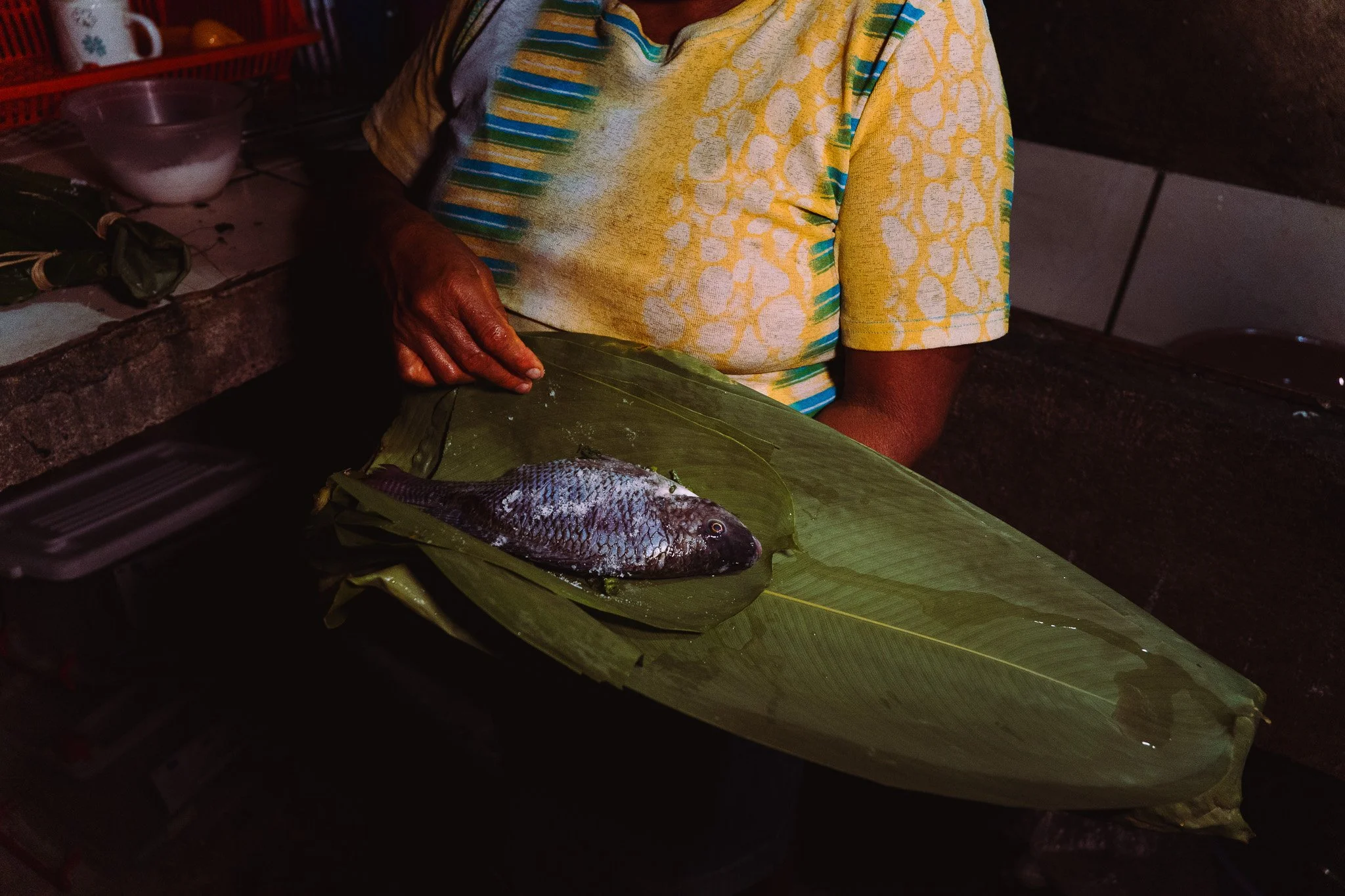 A person in a colorful yellow and blue patterned shirt preparing a fish wrapped in banana leaves on a kitchen countertop.
