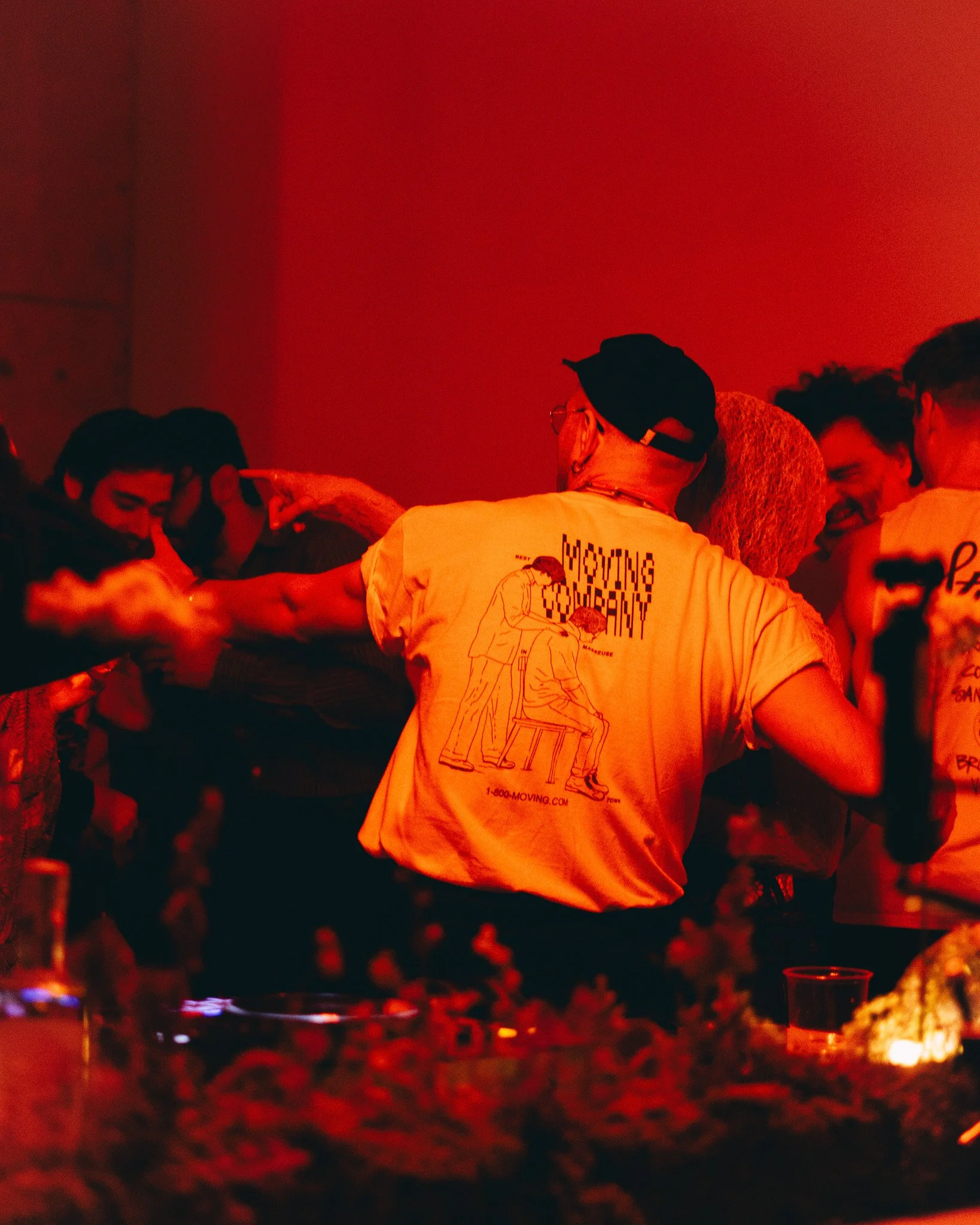 People at a nighttime gathering or party with red lighting, some sharing a contact lens, and a person wearing a white T-shirt with a cartoon graphic and text, surrounded by food, drinks, and candles.