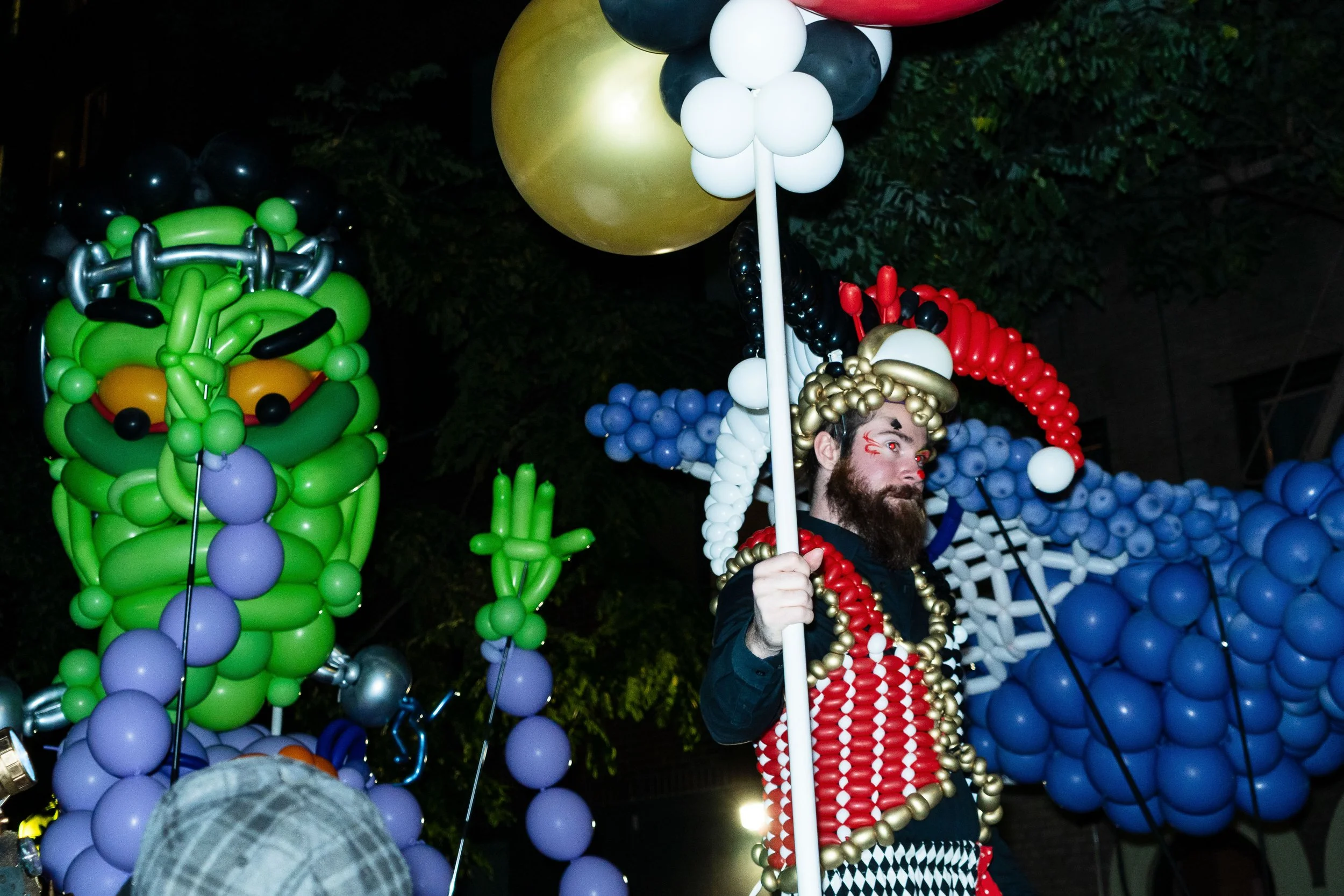 A man dressed in a costume made of colorful balloons, including a gold crown and beads, holding a white pole, with intricate balloon sculptures in the background, at a night event.