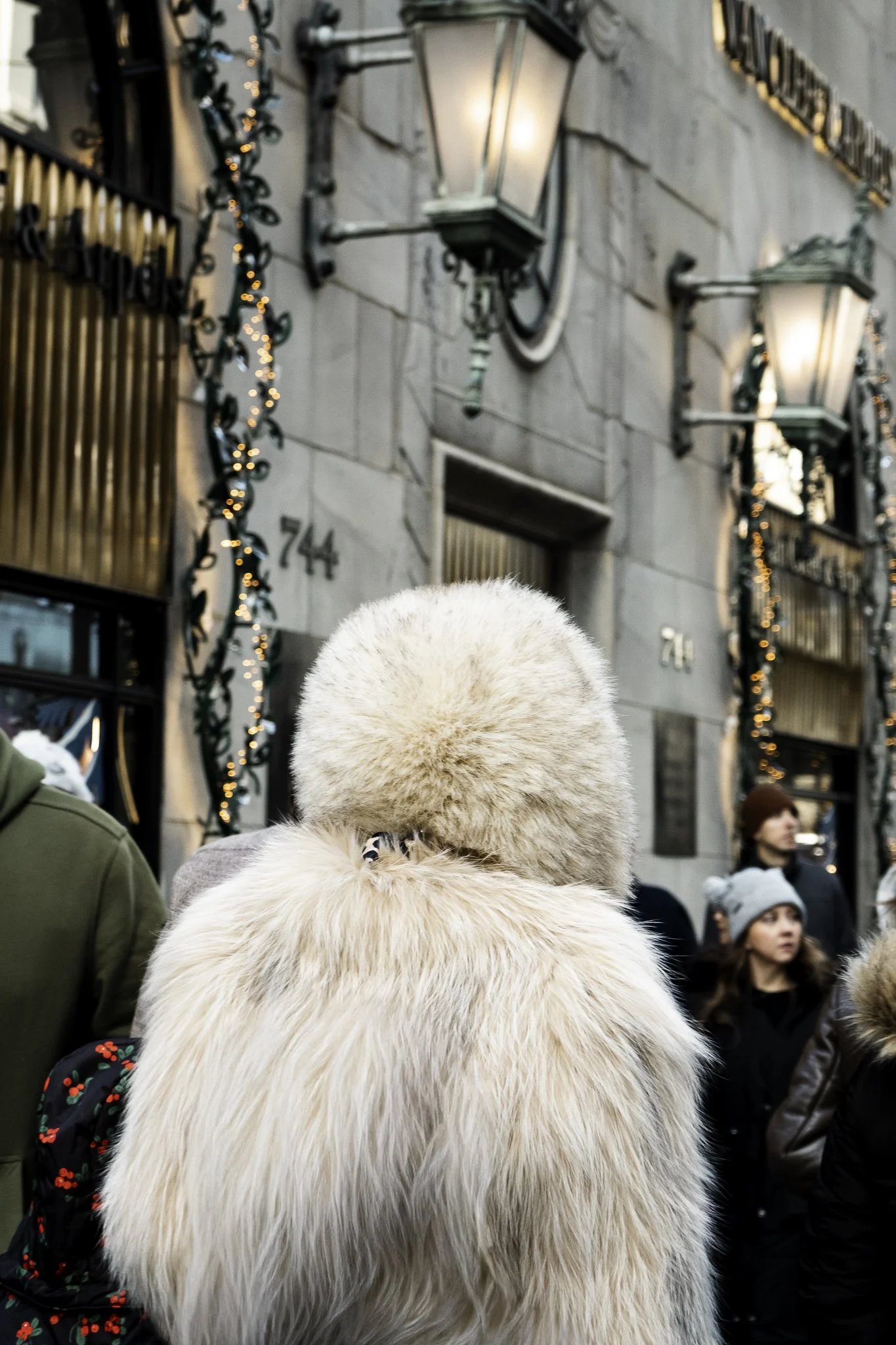 A person wearing a fluffy beige hat and coat standing among a crowd on a city street decorated with string lights, with buildings, a street lamp, and other pedestrians visible in the background.