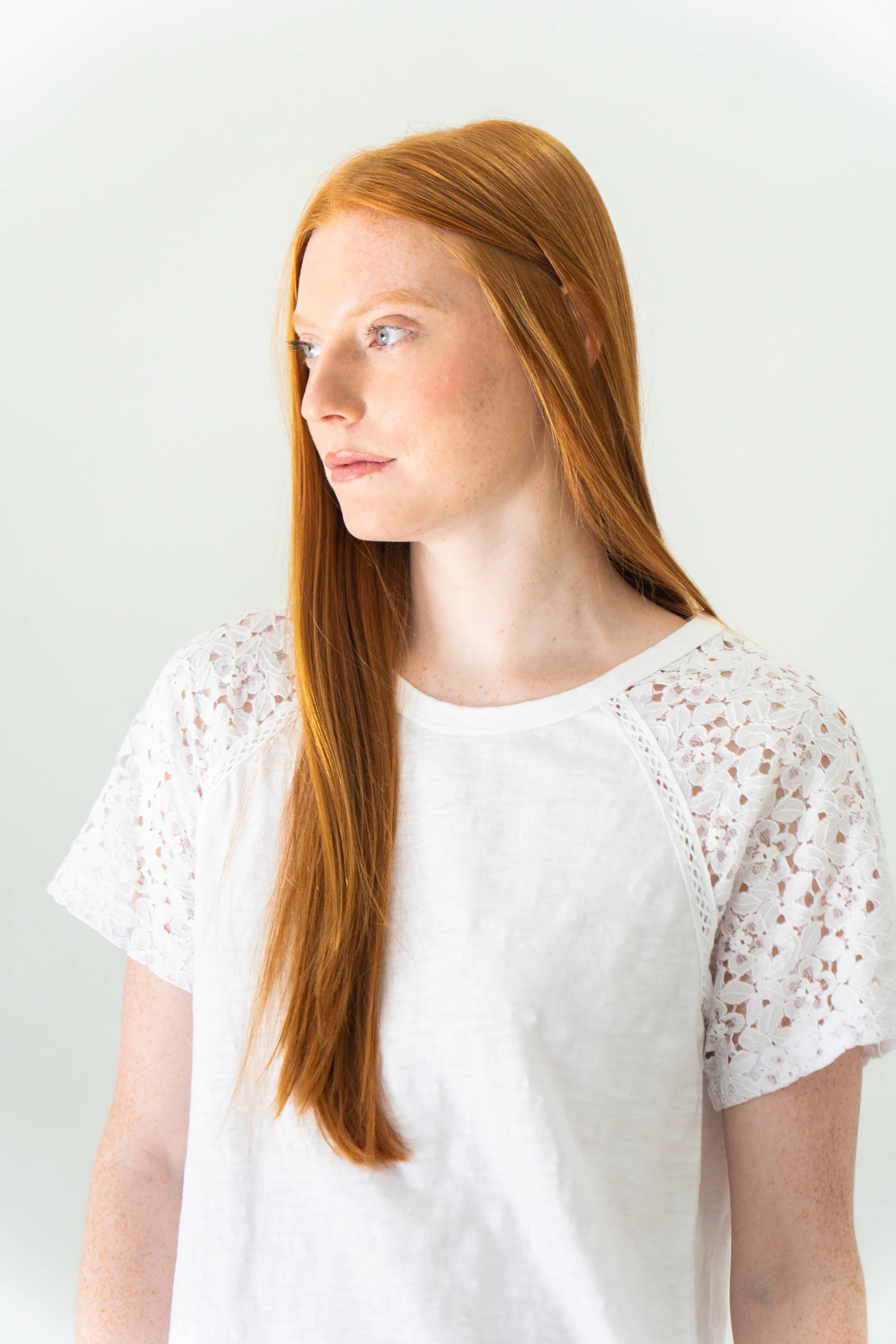 A young woman with long red hair wearing a white top with lace sleeves, looking to the left, standing against a plain light background.