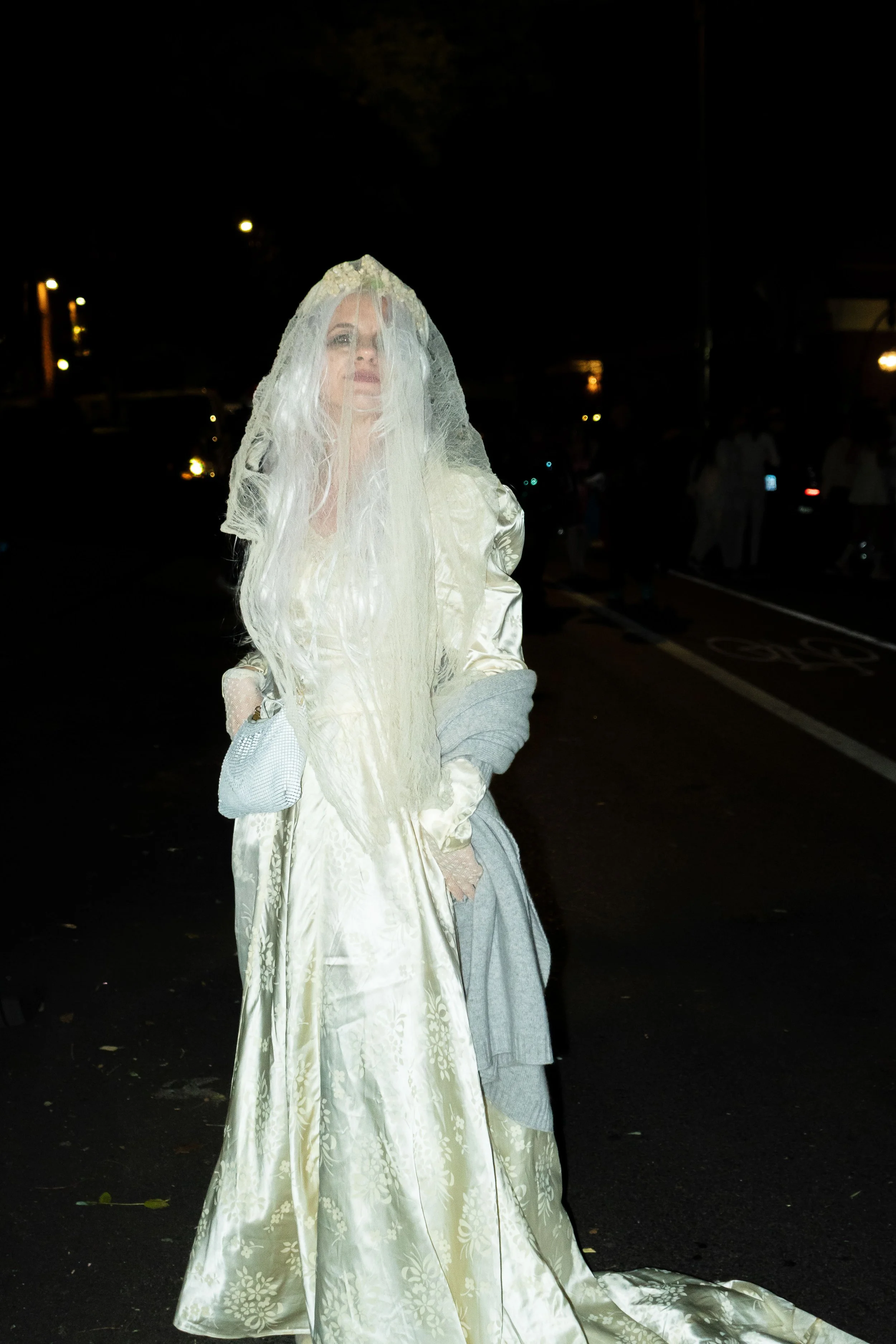 A woman dressed in a vintage, golden, floral-patterned satin bridal gown with a long train, wearing a lace veil, and holding a small silver handbag, standing outdoors at night.