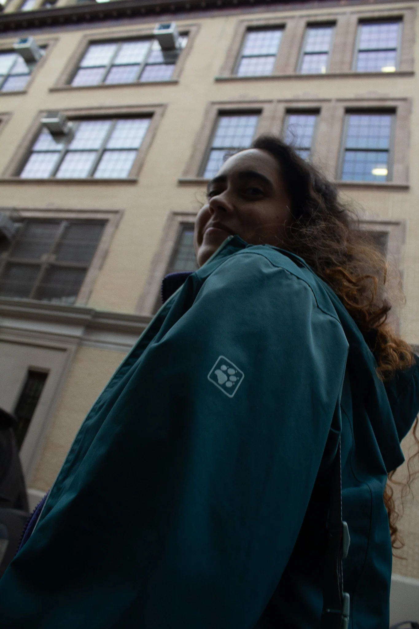 A person with curly hair wearing a teal jacket with a paw print logo on the sleeve, looking back at the camera, with a beige building with multiple windows in the background.