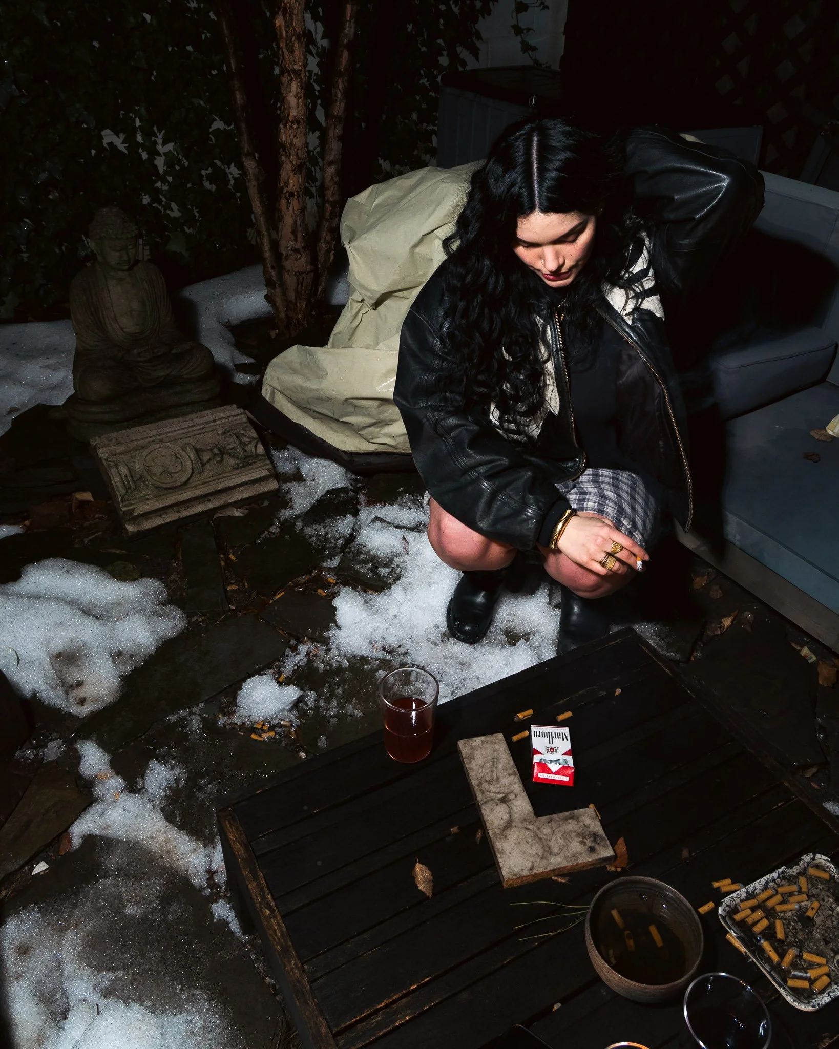 A woman with long black curly hair wearing a black leather jacket and plaid shorts crouching on a wooden patio at night, surrounded by snow and empty drink glasses, cigarette butts, a pack of cigarettes, and a cigarette ashtray, with a small Buddha s