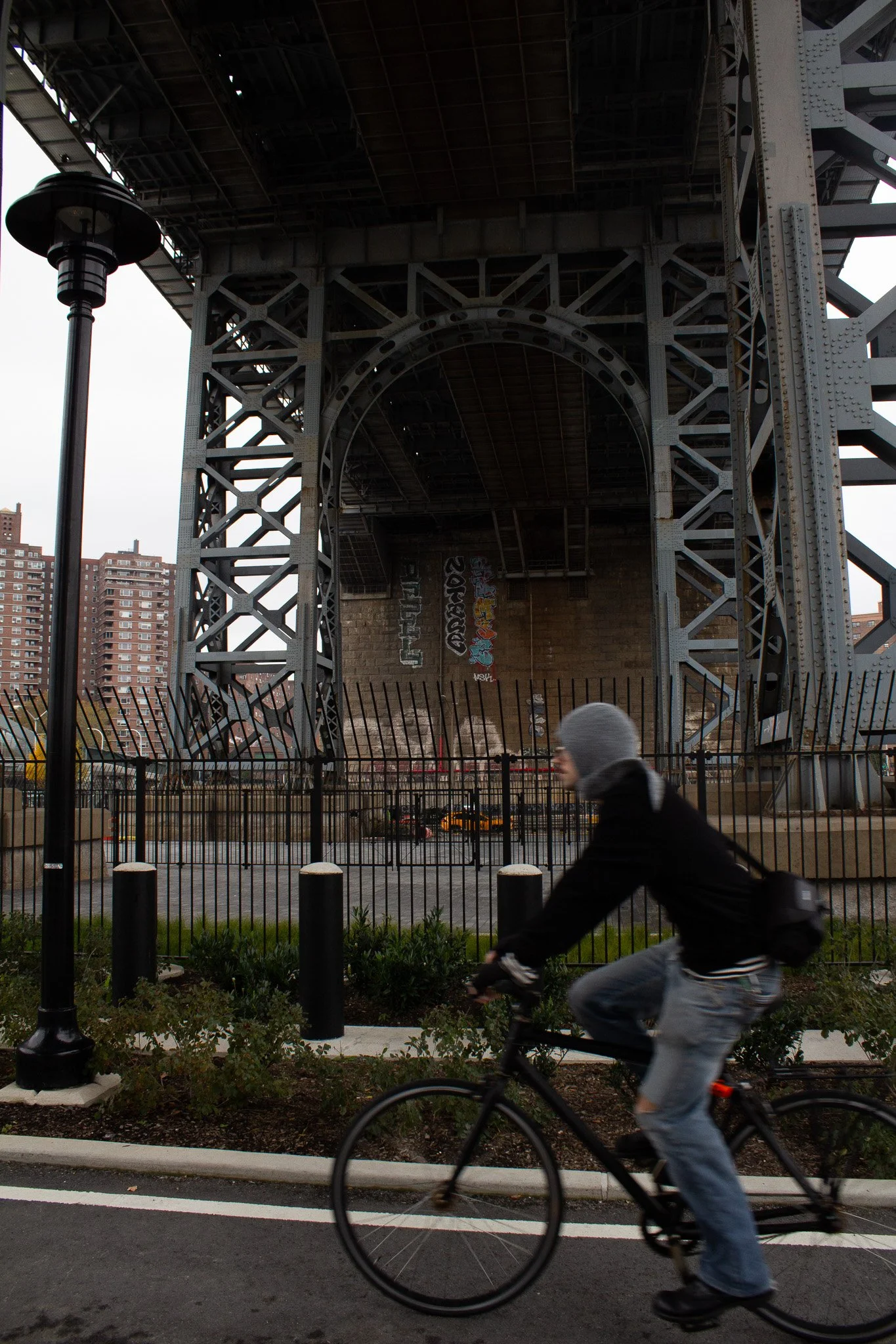 A cyclist riding past a large bridge with graffiti on the underside in an urban setting.