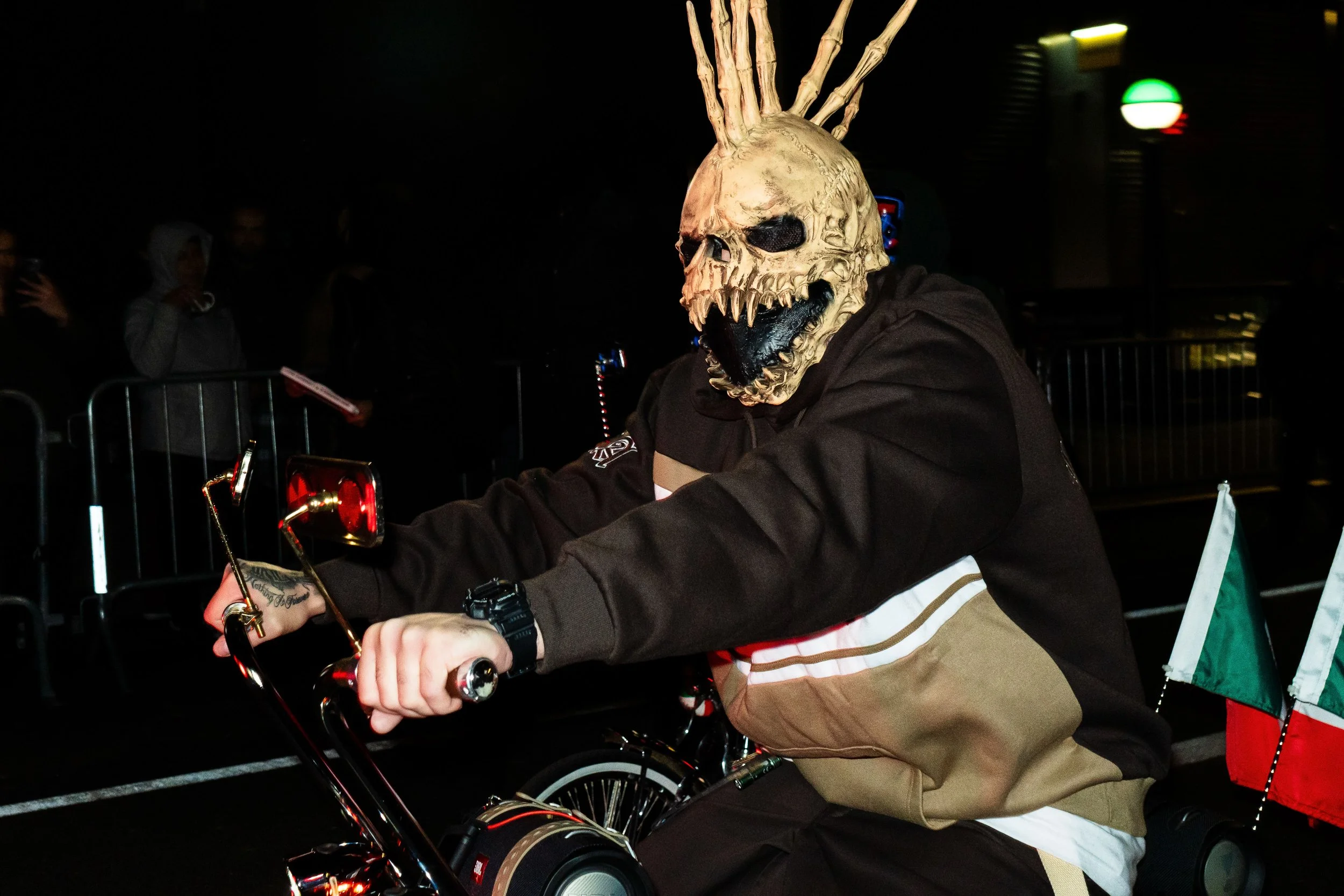 Person wearing a skull mask with spikes on head, riding a motorcycle at night, with Italian flags attached to the bike.