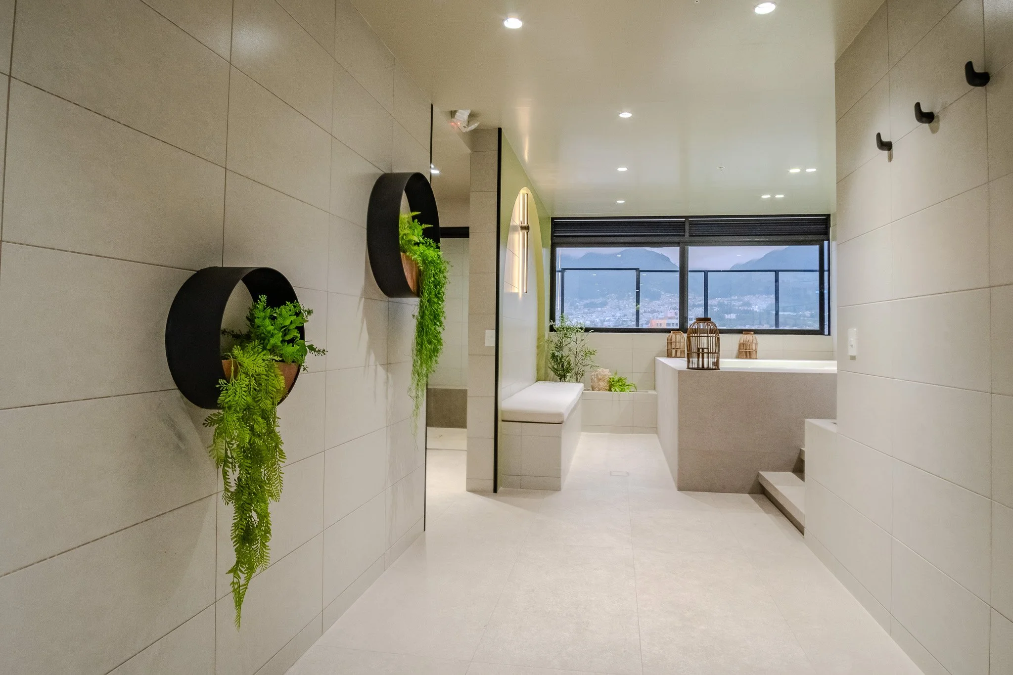 Modern interior with beige tiled walls and floors, black planters with green plants, a bench, and a large window showing a cityscape and mountains outside.