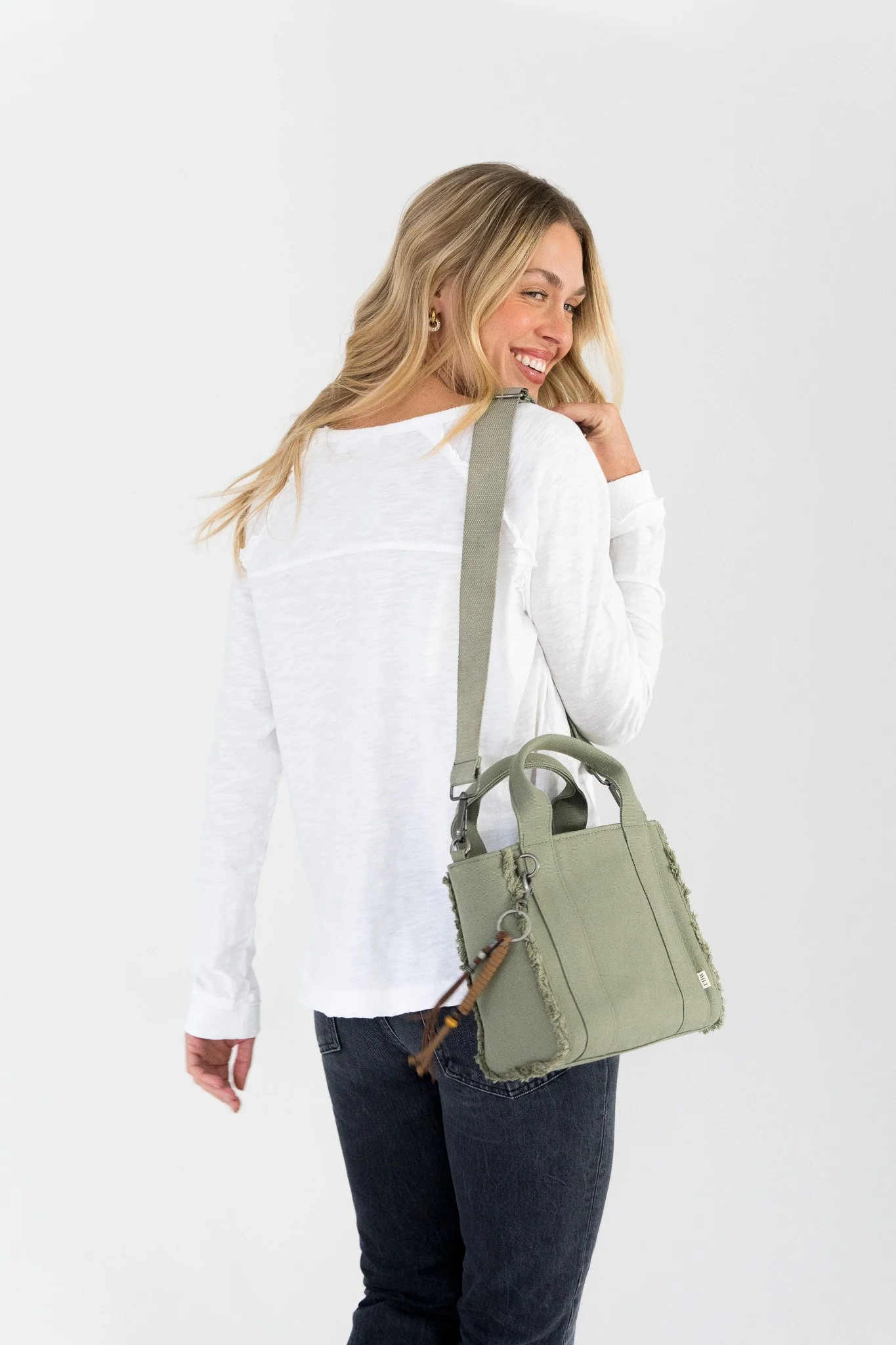 A woman with blonde hair, wearing a white long-sleeve shirt and dark jeans, carries a light green handbag over her shoulder, standing against a plain white background.