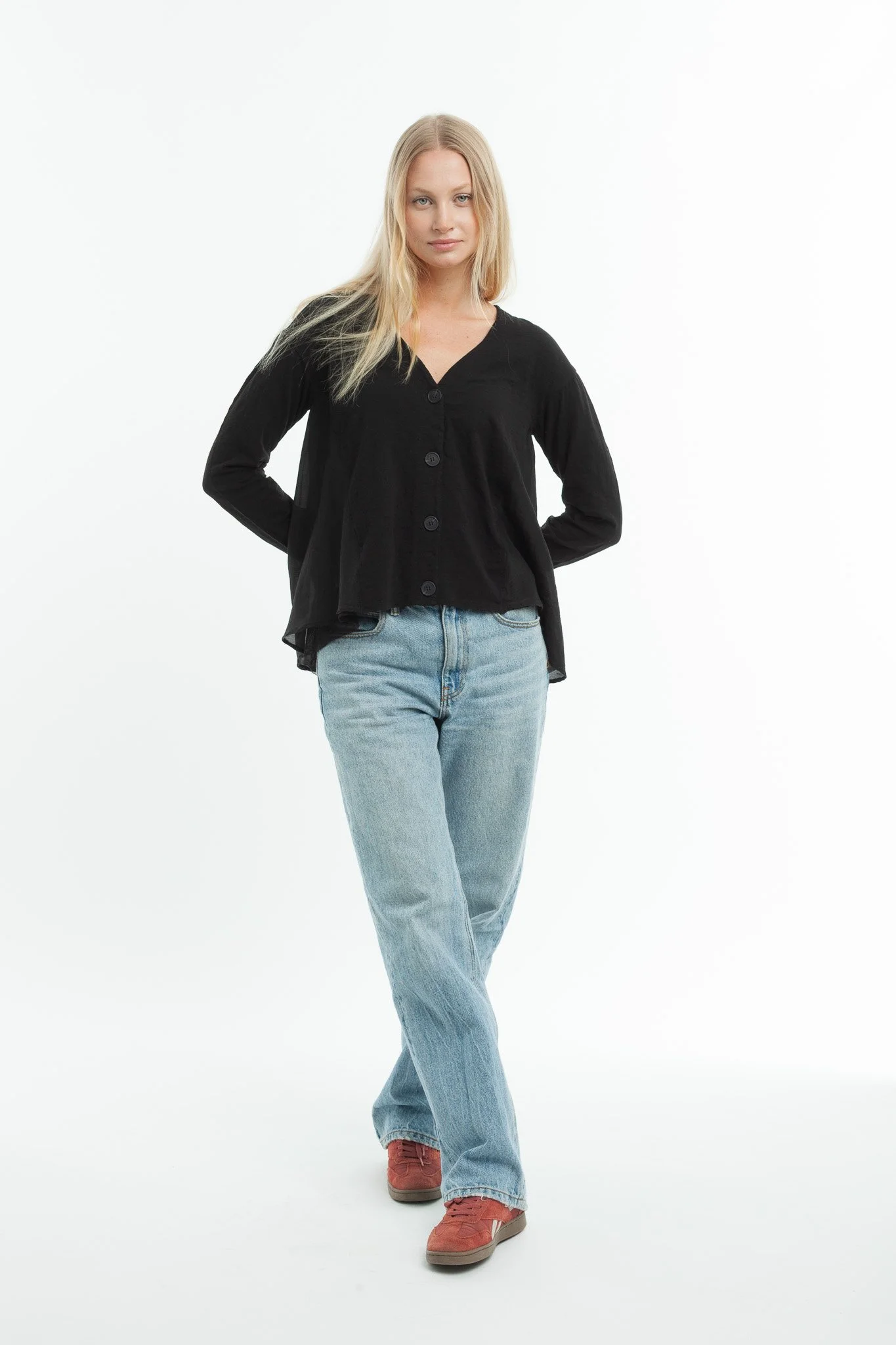 A woman with long blonde hair wearing a black button-up shirt, light blue jeans, and red shoes, standing against a plain white background.