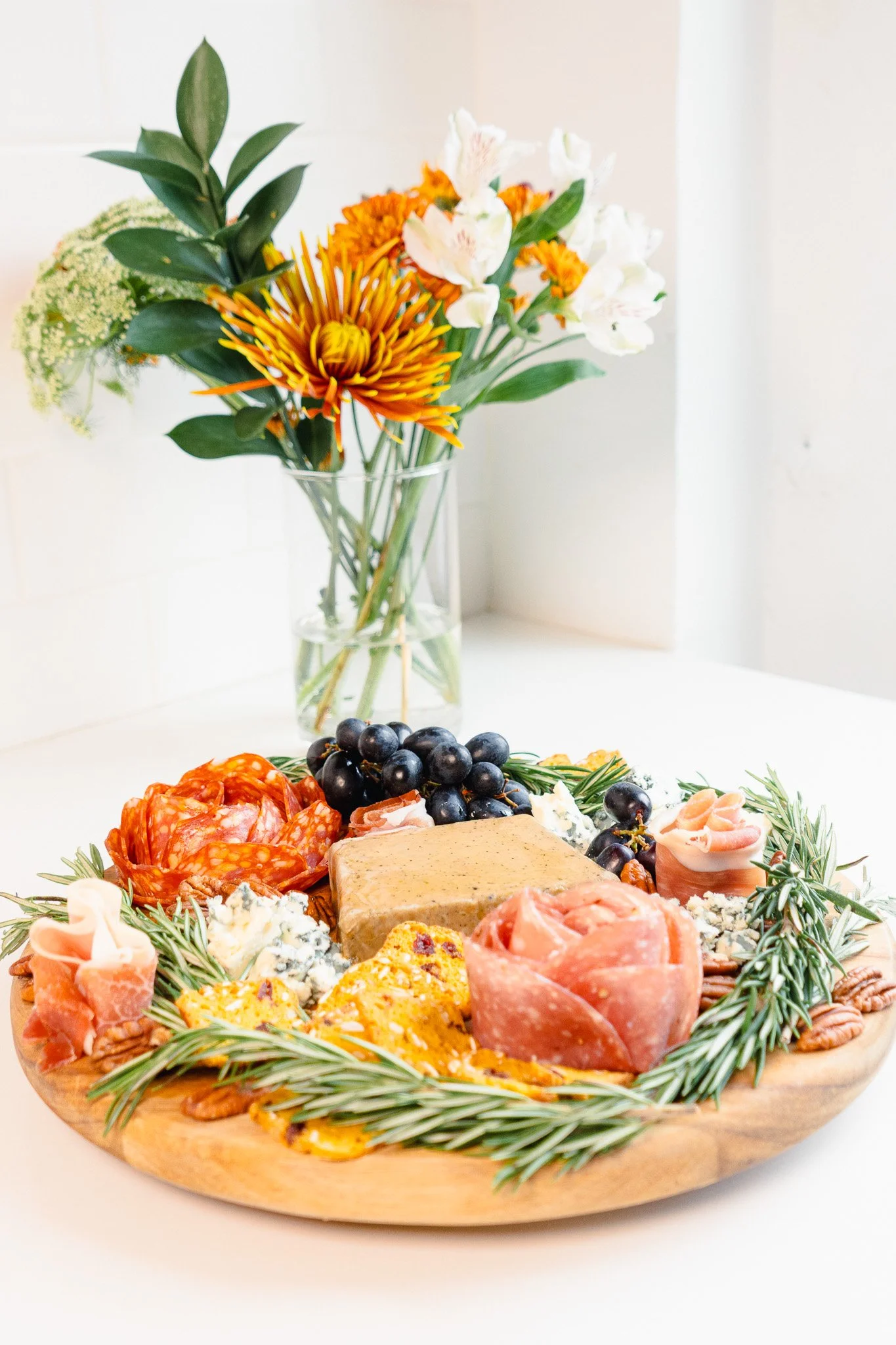 A wooden serving platter with assorted meats, cheeses, grapes, and herbs, surrounded by sprigs of rosemary, and a bouquet of white and orange flowers in a vase in the background.