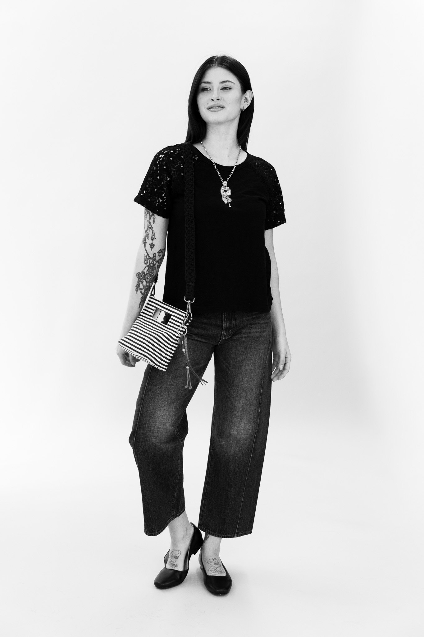 A woman with long dark hair wearing a black lace sleeve top, dark jeans, and black high heels. She has tattoos on her left arm and wears a necklace and a striped handbag on her shoulder. She is standing against a plain white background.