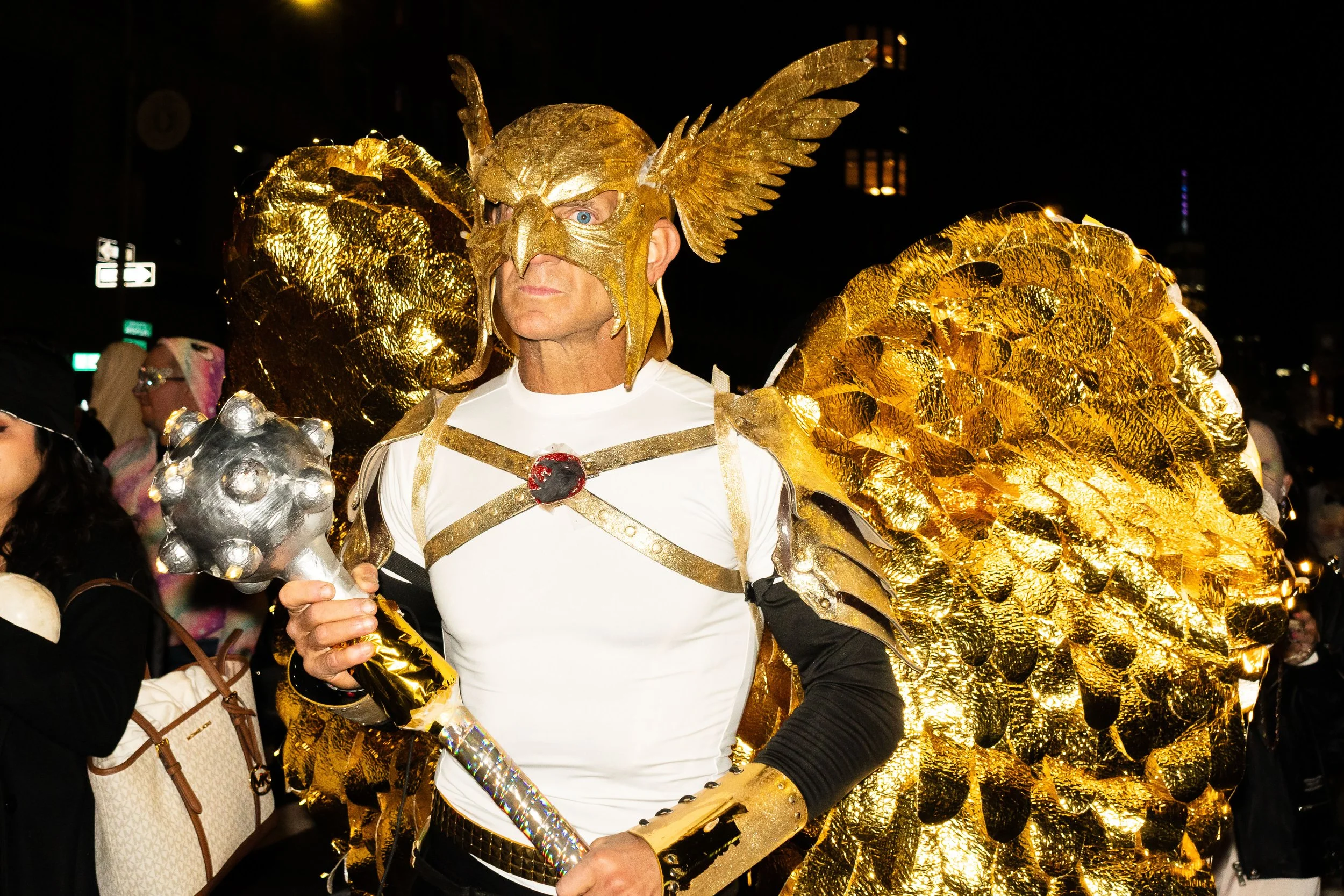 Person dressed in a gold and white costume resembling a mythological or fantasy character, holding a hammer, with large gold wings and an ornate helmet, participating in a nighttime event or parade.