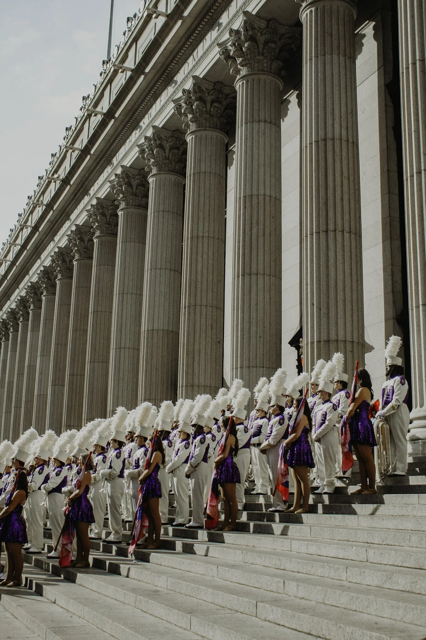 A group of people dressed in uniformed costumes, standing in formation on the steps of a neoclassical building with tall columns, participating in a parade or ceremony.
