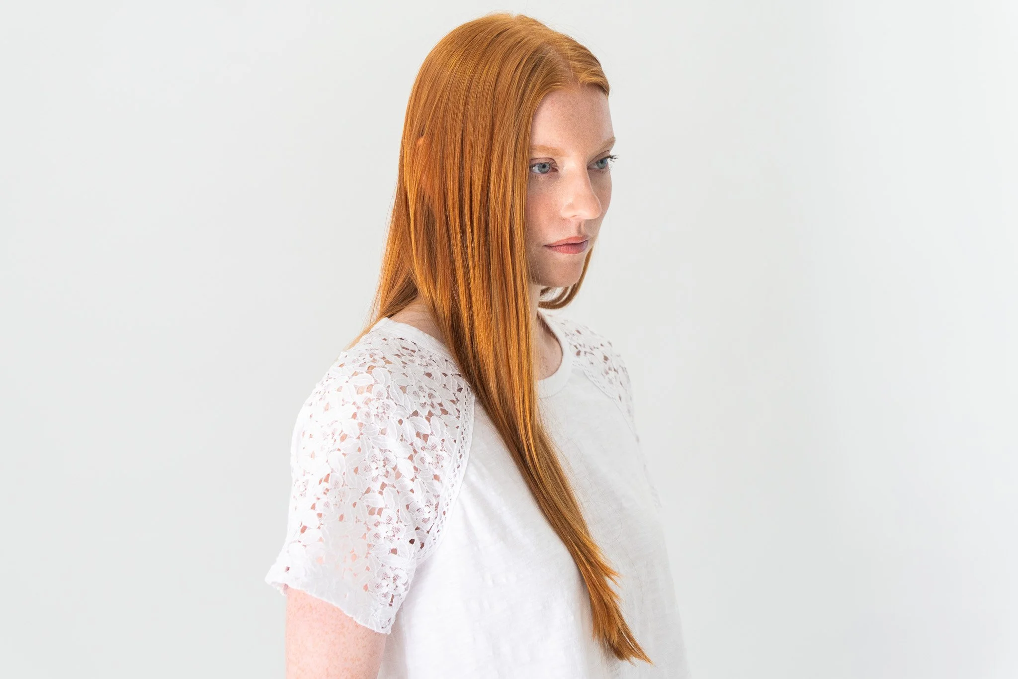 A woman with long red hair wearing a white lace top standing against a plain white background.