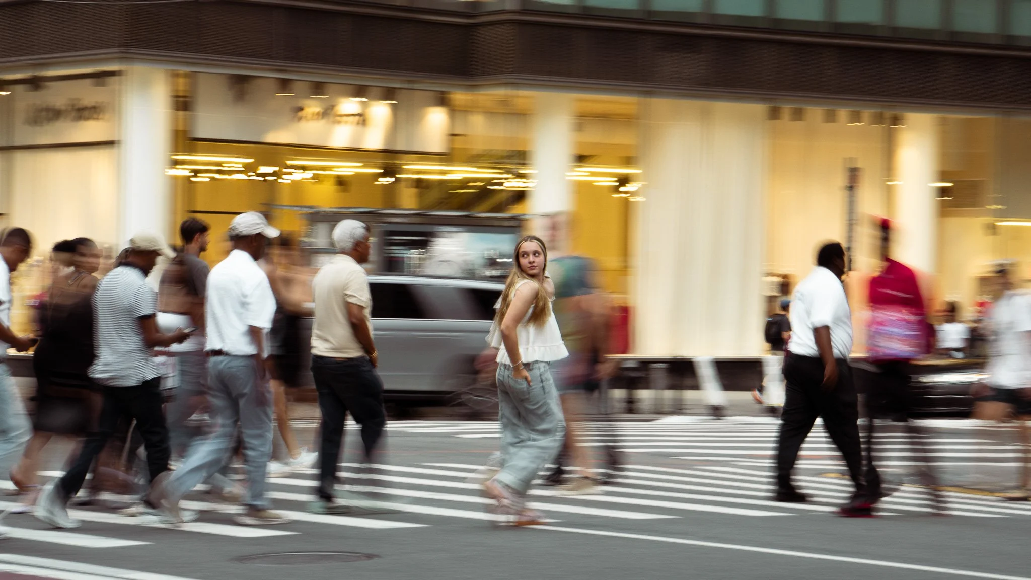People walking in crosswalk on city street at dusk, reflected lights from buildings, young woman with long blonde hair in white sleeveless top looking back.