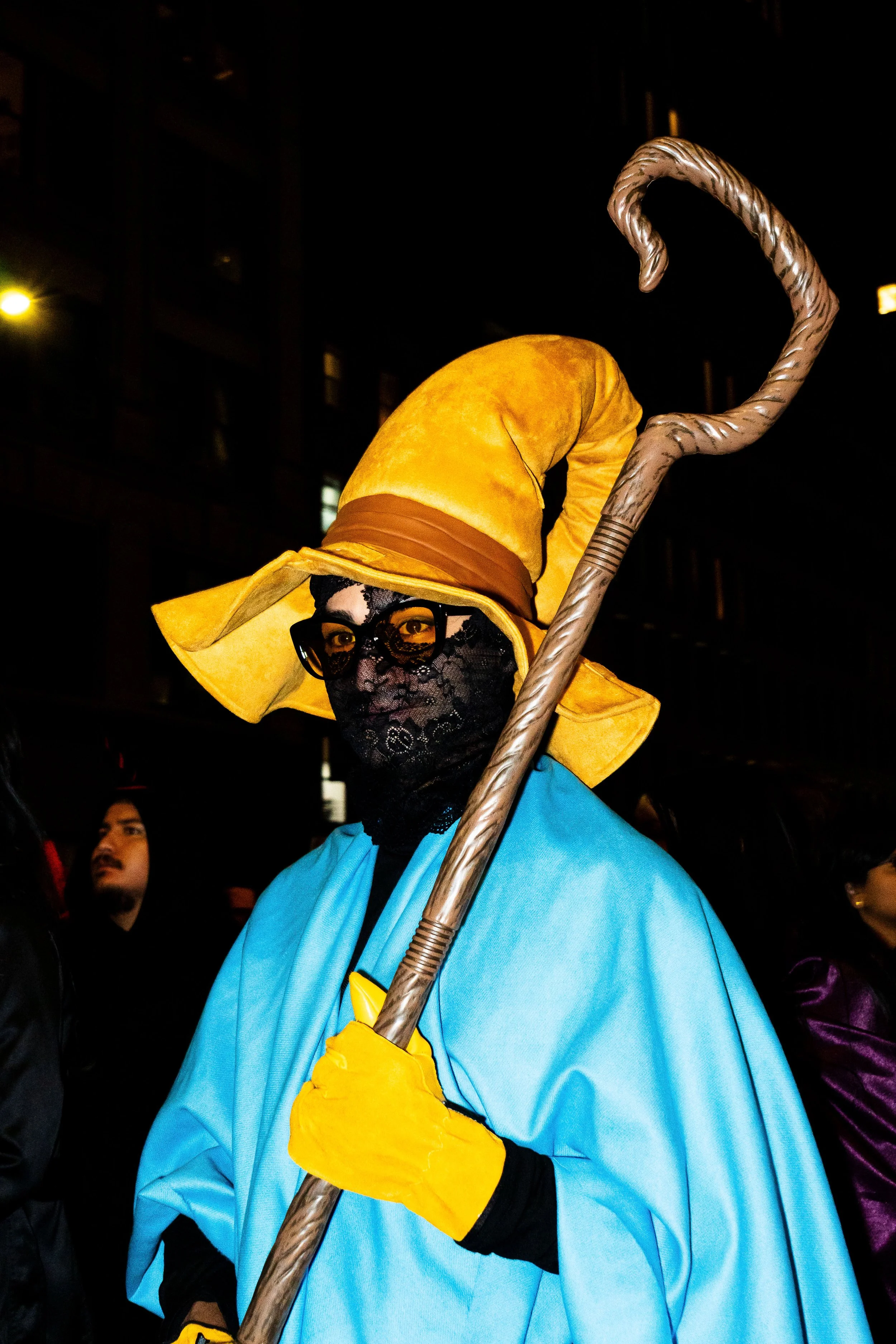 Person dressed as a wizard with a large yellow hat, black lace face mask, glasses, yellow gloves, and a blue robe, holding a staff, at night in a crowded outdoor setting.