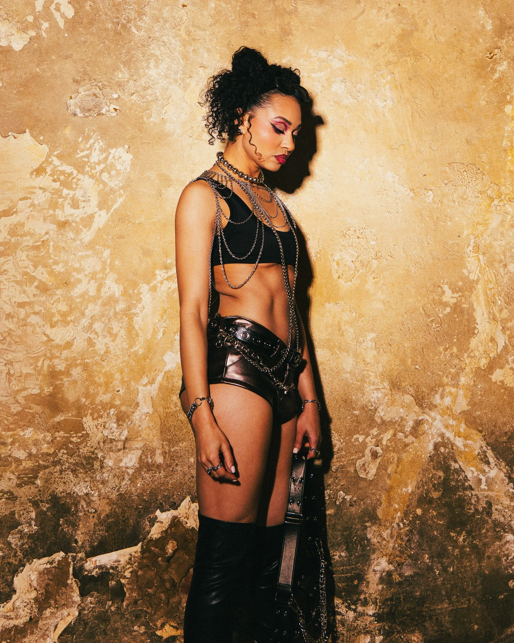A woman with dark, curly hair styled in a high bun, wearing dramatic pink and black eye makeup, and bold red lipstick. She is dressed in black leather shorts, a black top with cutouts, and layered silver chains, accessories, and jewelry. She stands a