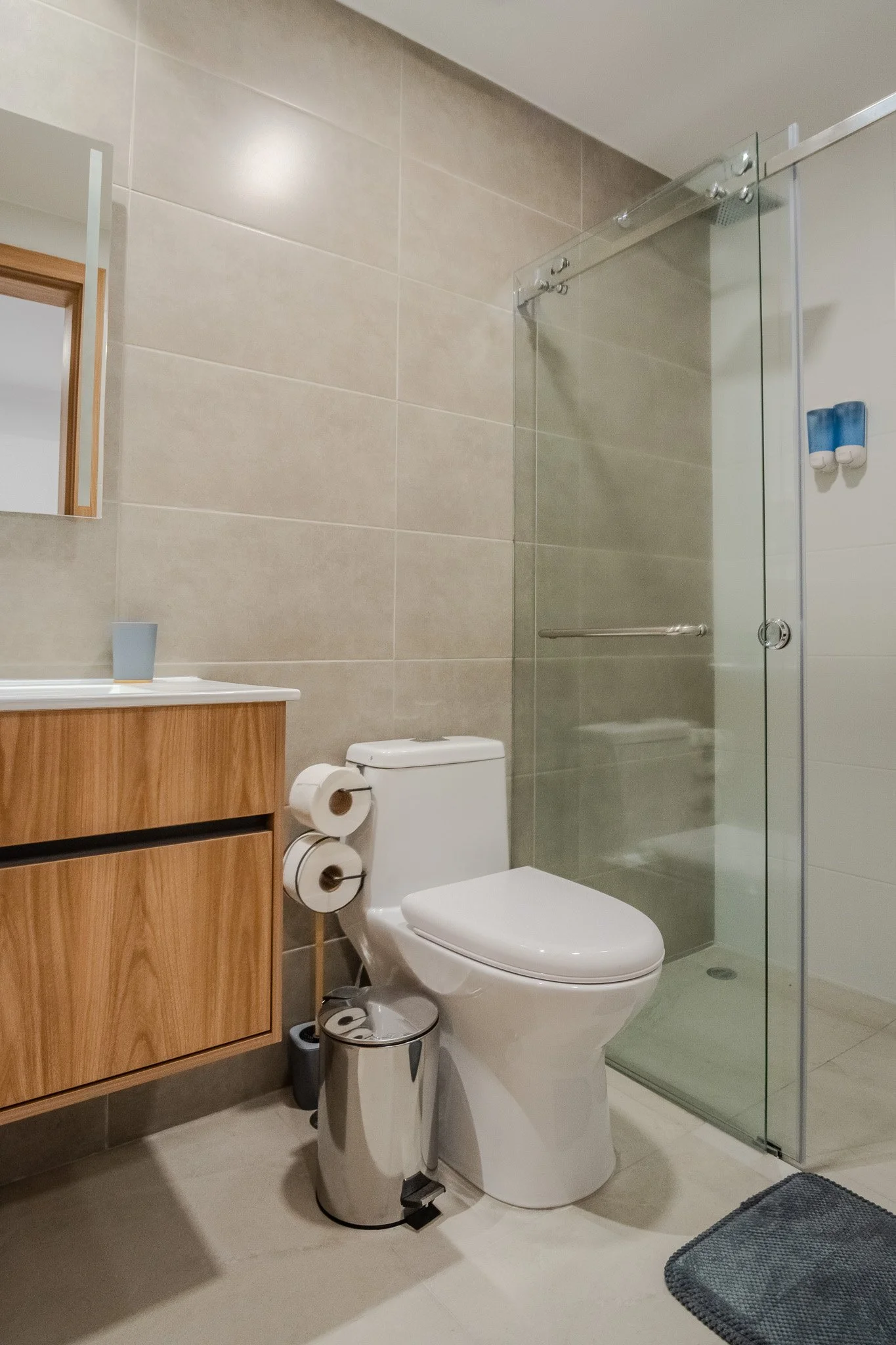Modern bathroom with beige tiled walls, a white toilet, a wooden vanity with a small mirror, and a glass shower enclosure. Toilet paper rolls are on a stand next to the toilet, and a small waste bin is nearby. A blue bath mat is on the floor.