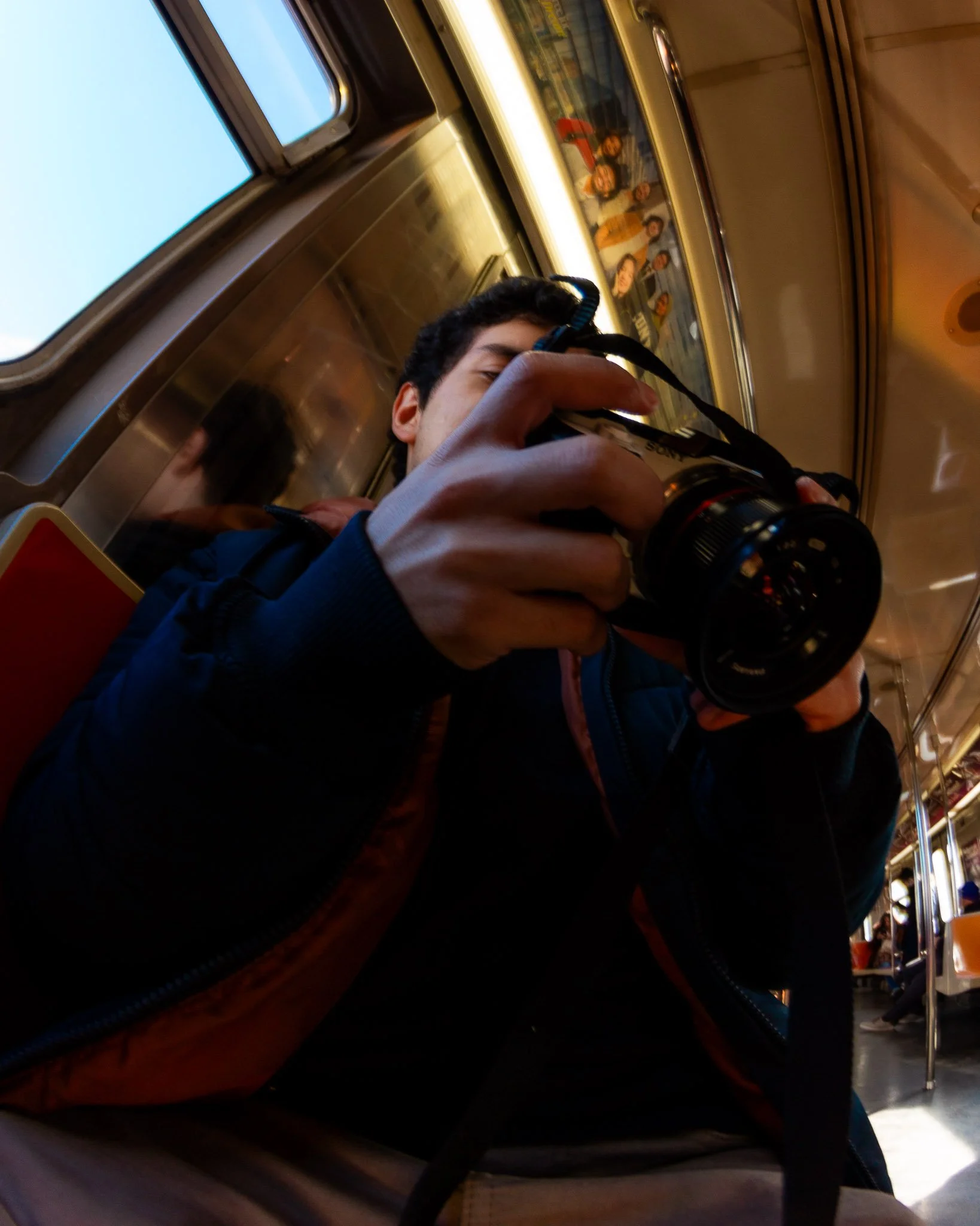 Person taking a photo with a camera inside a train, with a reflection of their face in the mirror and a window showing the blue sky.